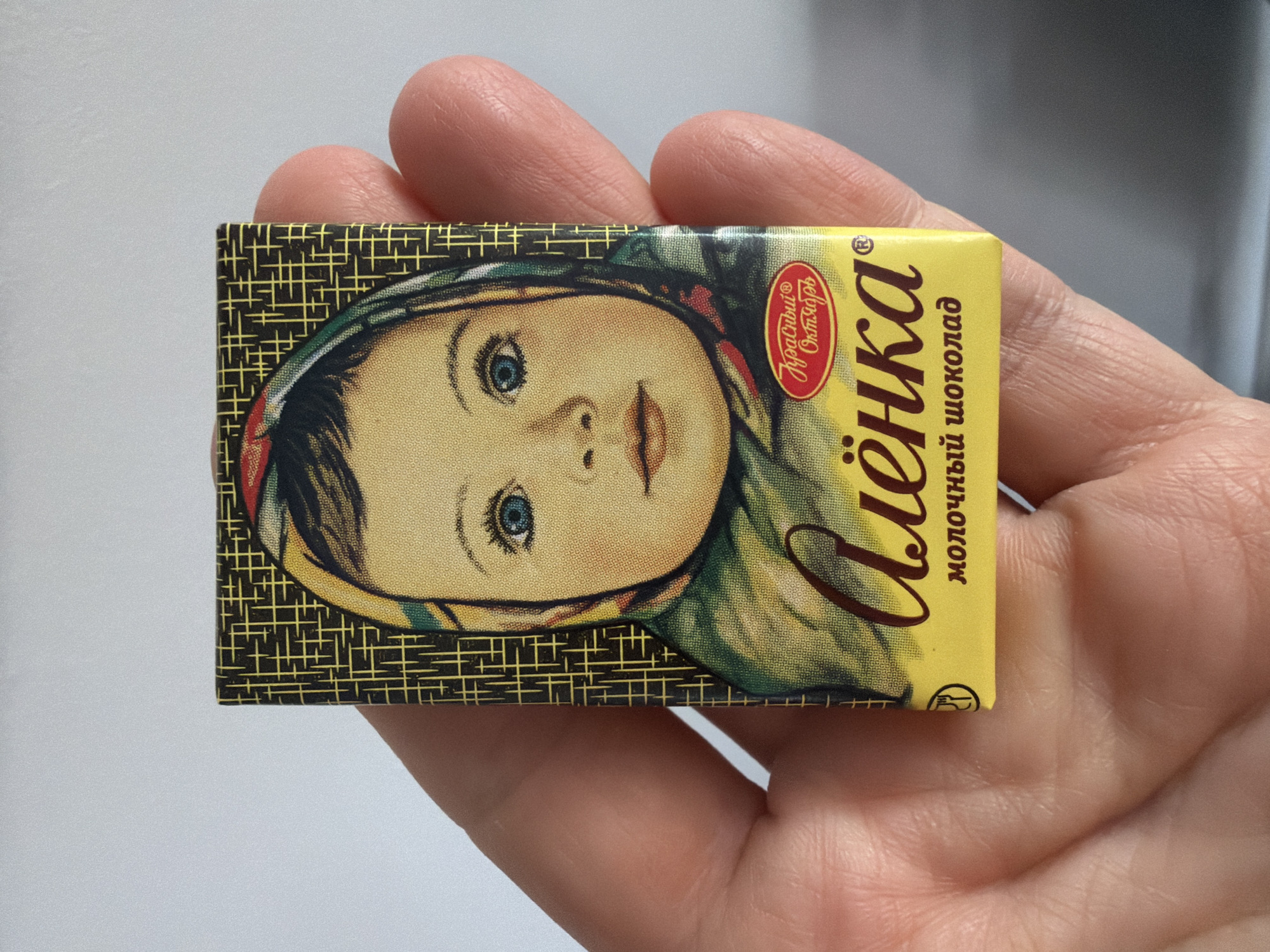 A hand holds a small rectangular chocolate bar wrapper. The wrapper has a yellow background on the right side, with the name "Alenka" in Cyrillic script written in brown cursive, followed by "молочный шоколад" (milk chocolate) in smaller letters. To the left of the text, a portrait of a young girl with blue eyes and a headscarf covers the remaining portion of the wrapper. The backdrop of the girl's portrait features a black background with a yellow grid pattern. On the top left of the yellow background of the wrapper, there is a logo in red, reading "Красный Октябрь" (Red October). The photo appears to be taken indoors, with a neutral-colored background, likely a wall or a tabletop. It is not possible to determine the location of the city of Chaikovsky, Russia based on the provided image.