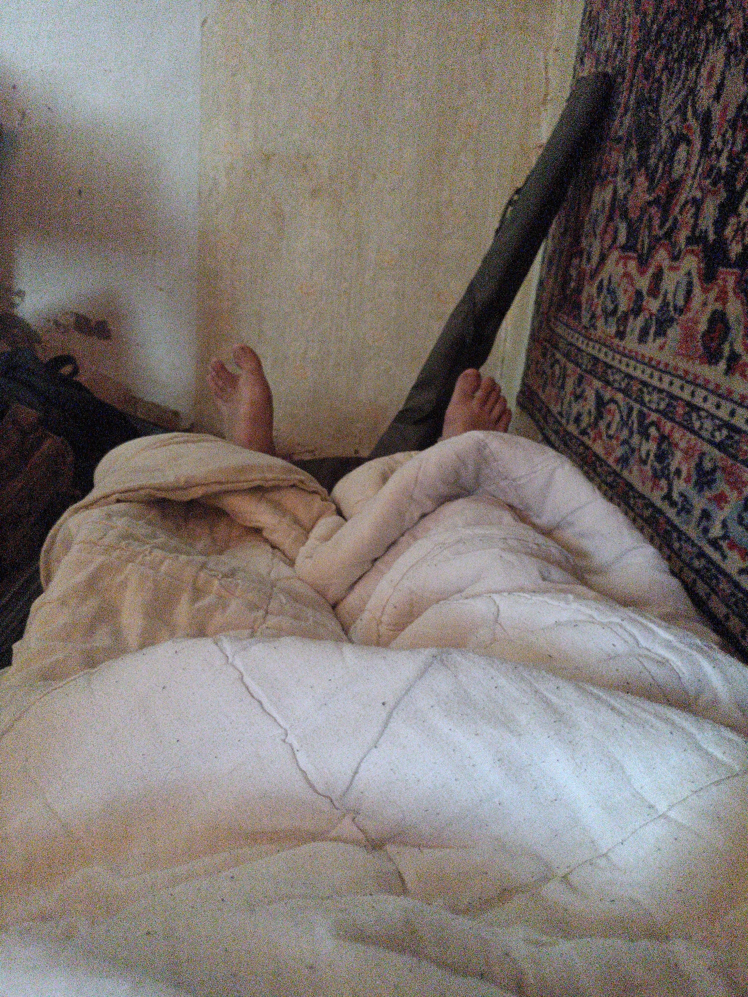 The image depicts a close-up view of a person's bare feet protruding from under a blanket in a dimly lit room. The feet, with visible toes, are positioned slightly upwards, suggesting the person is lying down. The blanket is a textured, light-colored fabric, folded and rumpled around the feet. In the background, a portion of a wall is visible, appearing textured and somewhat discolored. To the right, a dark, cylindrical object leans against the wall, and a section of a patterned textile, possibly a rug or tapestry with intricate designs in red, blue, and white, hangs on the wall. The overall impression is of a simple, possibly rustic, indoor setting. The image does not provide specific clues to pinpoint the location in Petro-Mykhailivka, Ukraine, beyond it being an interior space.