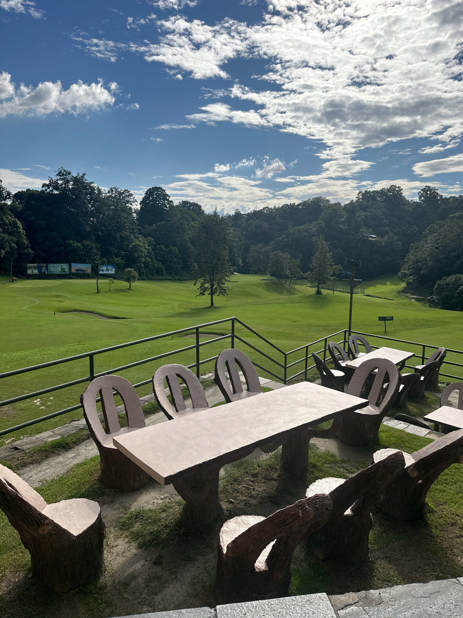 An outdoor seating area with concrete tables and chairs designed to resemble tree stumps and logs overlooks a golf course in Kageshwori Manohara, Nepal. The seating area, positioned on a grassy slope with a metal railing, features several tables and chairs. In the background, a lush green golf course stretches out, bordered by dense trees on rolling hills. The sky above is blue with scattered white clouds, indicating fair weather. The scene is devoid of people.