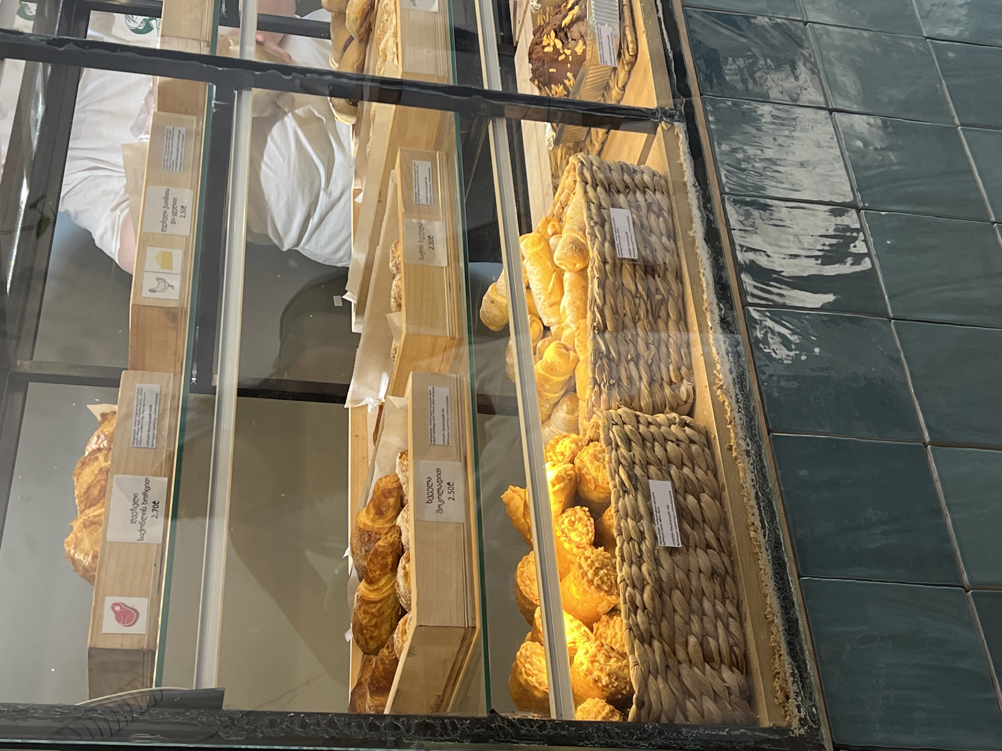 The image shows a display case at a bakery in Batumi, Georgia. The display case contains various baked goods, including pastries and bread. The baked goods are arranged in wooden crates and woven baskets. Some of the items have labels with Georgian text and prices. In the background, a person wearing a white t-shirt can be seen behind the counter. The bakery has a modern interior with tiled walls and glass display cases. The lighting in the display case is warm and inviting, highlighting the texture and color of the baked goods.