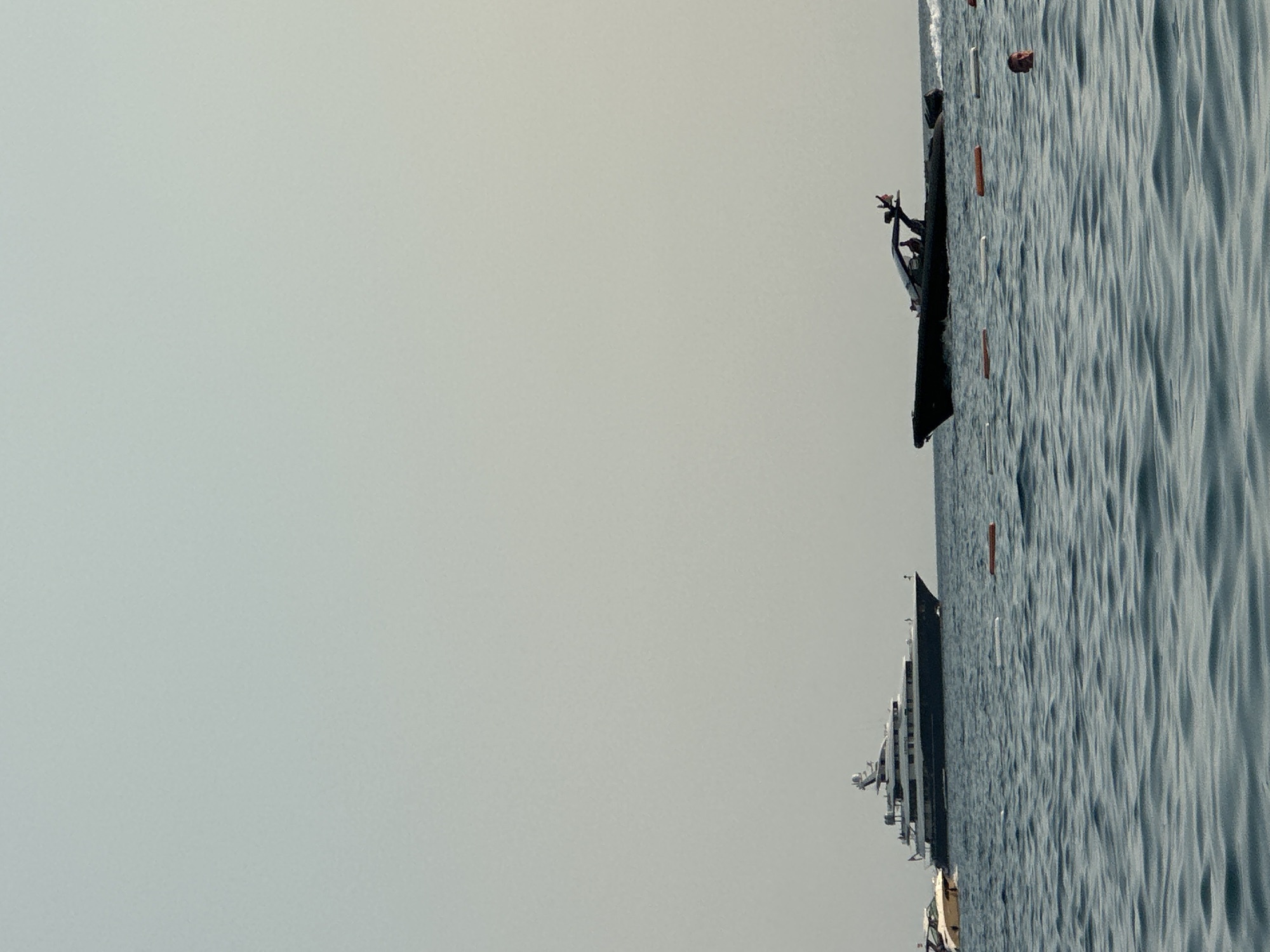 In the waters off Boreti, Montenegro, a large, dark-hulled yacht is anchored in the distance, its multiple decks and superstructure clearly visible against a pale sky. Closer to the foreground, a sleek, black speedboat with several occupants cuts through the waves, leaving a visible wake. The water is choppy, with a series of orange and white buoys marking a swimming area. In the mid-ground, a man's head is visible above the water, looking towards the viewer. The scene suggests a leisure activity in a coastal area.