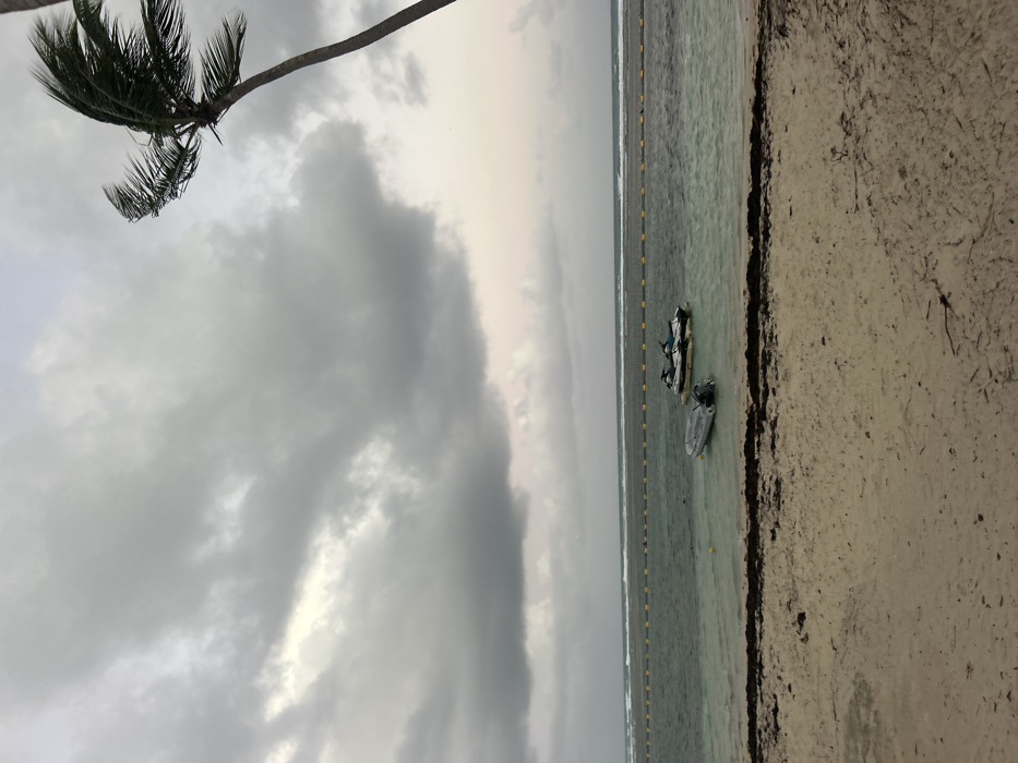 The image presents an overcast coastal scene located in Sainte-Anne, France. The upper two-thirds of the frame are dominated by a sky filled with layered dark grey and lighter grey clouds, showing a slightly brighter tone towards the upper right. The horizon separates the sky from a body of water, which transitions from a light greenish-blue near the shore to a darker blue further out, displaying small waves on the distant surface. A continuous yellow floating barrier extends horizontally across the mid-ground of the water. Positioned within the water, near this barrier, are three watercraft: two white and dark-colored jet skis are clustered together on the right, and a single grey inflatable boat equipped with an outboard motor floats to their left. A solitary yellow buoy is visible in the water near the inflatable boat. The foreground comprises a sandy beach of light beige color, featuring scattered dark brown seaweed and debris along the waterline. A section of a palm tree trunk, with its fronds visibly bent towards the left, indicating wind, is present along the extreme right edge of the frame. No individuals are visible within the scene.
