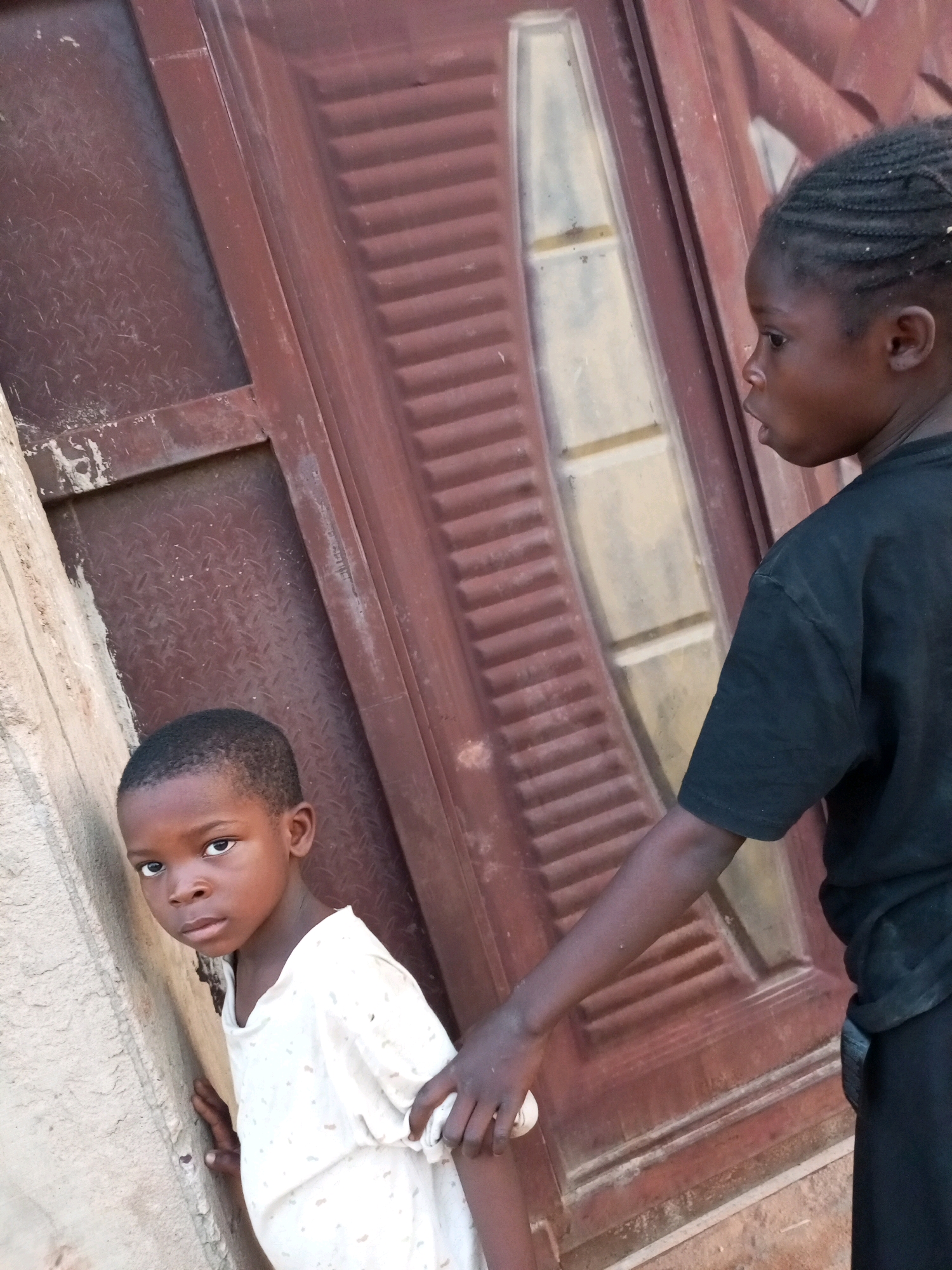 In Tempure, Nigeria, two young children stand against a weathered, reddish-brown door. The child on the left, facing forward, is wearing a light-colored, patterned t-shirt and has short, dark hair. Their gaze is directed at the camera with a serious expression. To the right, a second child, partially obscured, is seen from the side, looking away from the camera. This child has dark, braided hair and is wearing a black t-shirt. Their right arm is extended, with their hand near the younger child's shoulder, suggesting a possible interaction or gesture of guidance. The background consists of a textured, concrete wall to the left and the segmented surface of the door, which appears to have a matte finish with some signs of wear and tear. The lighting suggests it is daytime, with shadows indicating the direction of the light source.