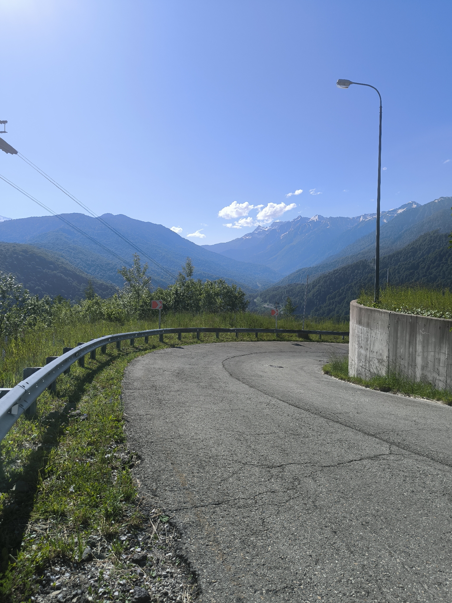 A paved road curves through a mountainous landscape under a clear blue sky. The road is bordered by a metal guardrail on the left and a concrete retaining wall topped with vegetation on the right. In the background, lush green mountains rise, with snow-capped peaks visible in the distance. A single street lamp stands tall on the right side of the road, and power lines traverse the upper portion of the image. A red and white directional sign is visible on the left side of the road, indicating a sharp turn. The overall scene suggests a scenic mountain road, likely in a resort area given the presence of a street lamp and guardrails. The location is identified as Estosadok, Russia.
