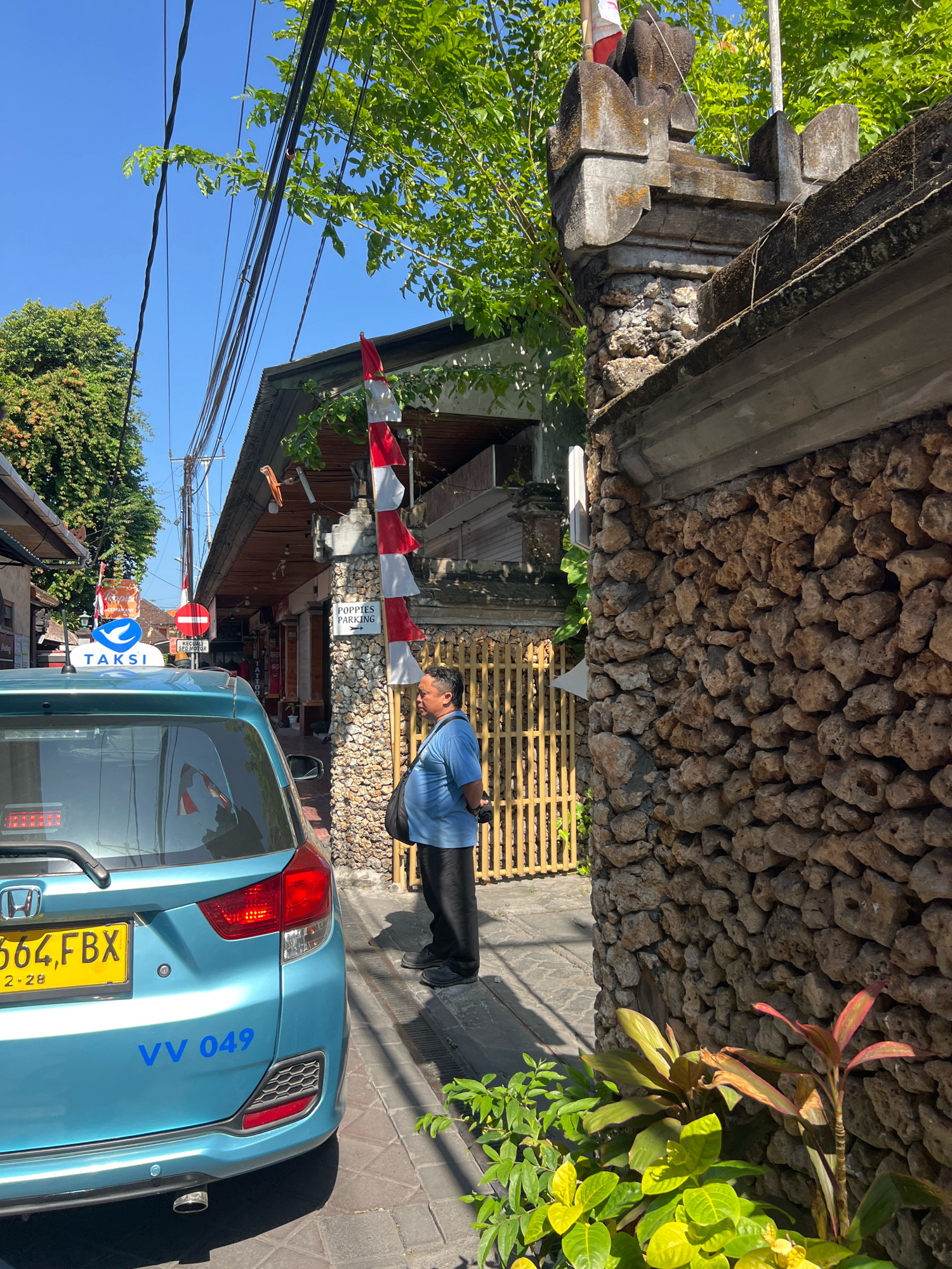 In Kuta, Indonesia, a light blue taxi is parked on a paved street under a clear blue sky. The taxi displays a yellow license plate with the numbers "664 FBX" and the date "12 . 28". Blue lettering on the rear of the car reads "VV 049". A man wearing a blue polo shirt and dark pants stands beside the taxi, facing away from the camera. He has a dark bag slung over his shoulder. In the background, a building with a weathered, rustic facade is visible, adorned with red and white flags. A sign on the building reads "POPIES PARKING". A stone wall with a wooden fence runs along the right side of the image, bordering lush green foliage. Overhead, a tangle of electrical wires crisscrosses the scene, partially shaded by the vibrant green leaves of trees.