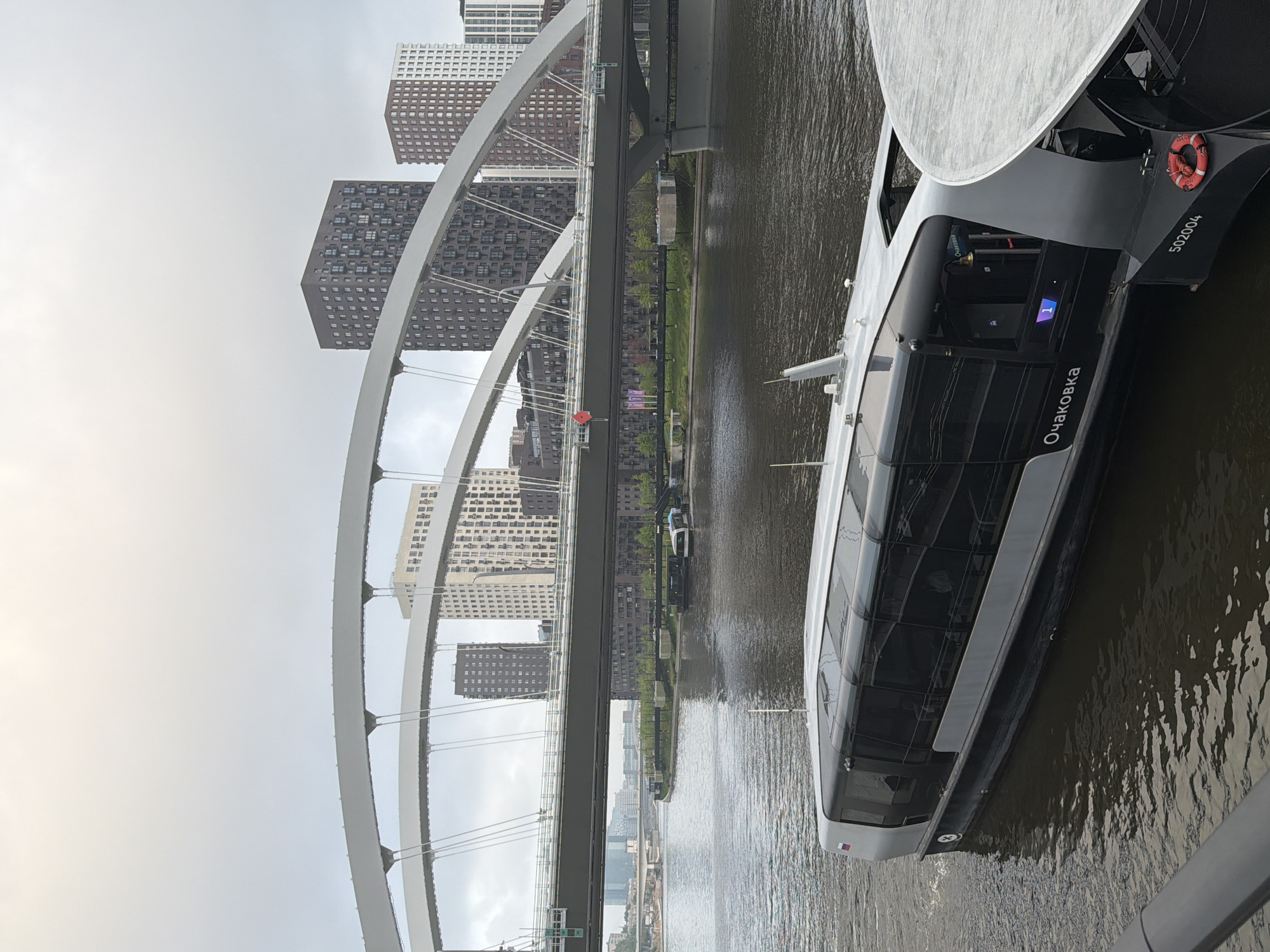 The image shows a river scene in Moscow, Russia, with the Zvenigorodskoe highway bridge in the background. A modern river vessel labeled "Очаковка" (Ochakovka) is docked along the riverbank. The vessel is gray and white with large windows and a life preserver visible at the rear. In the background, modern high-rise buildings are visible behind the bridge. The river appears calm, reflecting the overcast sky. There are some trees along the river bank, and the overall scene suggests an urban environment.