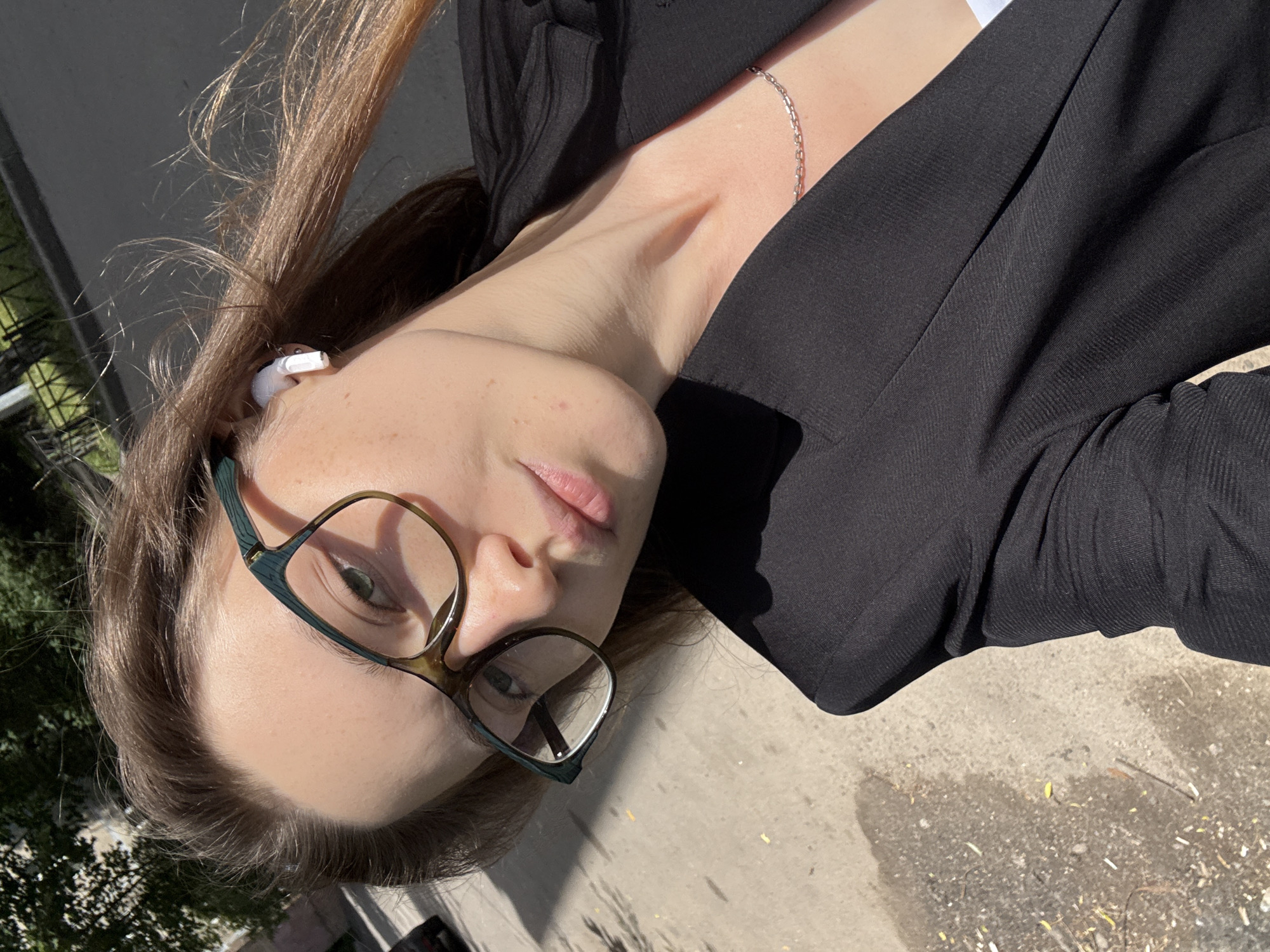 A young woman, wearing a black blazer and glasses, is captured in a close-up, upward-angle selfie. She has long, light brown hair that frames her face and a white earbud in her left ear. A delicate silver chain necklace is visible around her neck. Her expression is neutral, with her lips slightly parted. The lighting suggests it is daytime, with sunlight casting shadows. The background shows a textured, greyish surface, possibly pavement, with some scattered debris. The overall setting appears to be outdoors, with hints of greenery and what might be buildings in the distance. The image is oriented sideways.