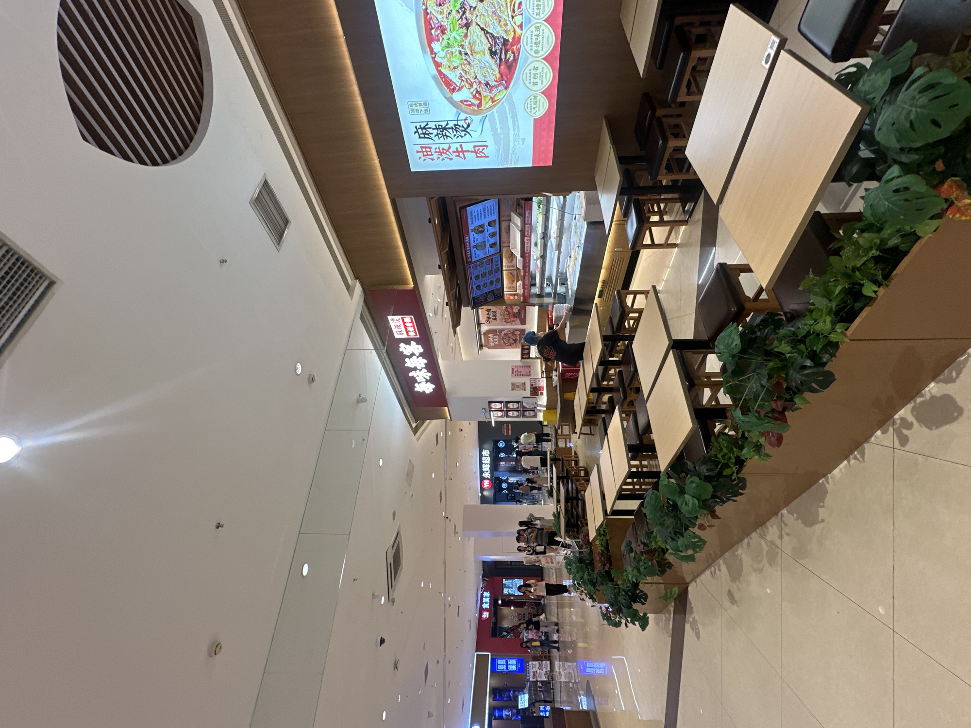 The image depicts an indoor dining area of a restaurant, likely in a commercial space like a mall or food court, located in the Chongqing High-tech Zone, China. The restaurant features a modern, clean aesthetic with light-colored tables and dark stools, arranged in a grid. A prominent digital menu board displays food items, and an illuminated advertising screen showcases a large image of a dish, likely the restaurant's specialty, with Chinese text. Behind the counter, a staff member wearing a blue hairnet and apron is visible, seemingly preparing or serving food. The floor is tiled, and decorative planters with lush green foliage are interspersed among the seating arrangements, creating a subtle natural element. In the background, other shopfronts and a general walkway of the commercial area are visible, indicating a bustling environment. The ceiling is modern, with integrated lighting and ventilation systems. The overall scene suggests a typical, contemporary fast-casual dining establishment.