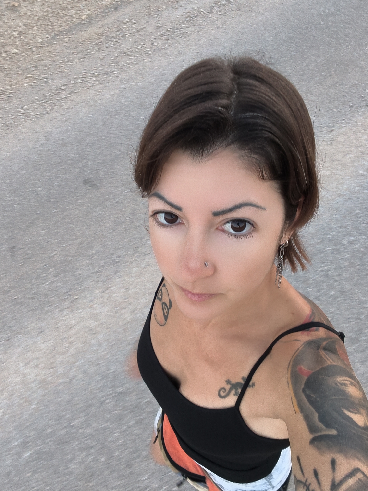 A high-angle, close-up, point-of-view shot shows a woman from the chest up, outdoors. She has short, dark brown hair and is looking directly at the camera. She is wearing a black tank top, and white shorts with an orange waistband is visible. She has tattoos on her left shoulder and chest, and a silver chain earring in her left ear. A silver nose stud is visible on her right nostril. The background is a rough, grey, asphalt surface. The image appears to have been taken in Lachish Regional Council, Israel.