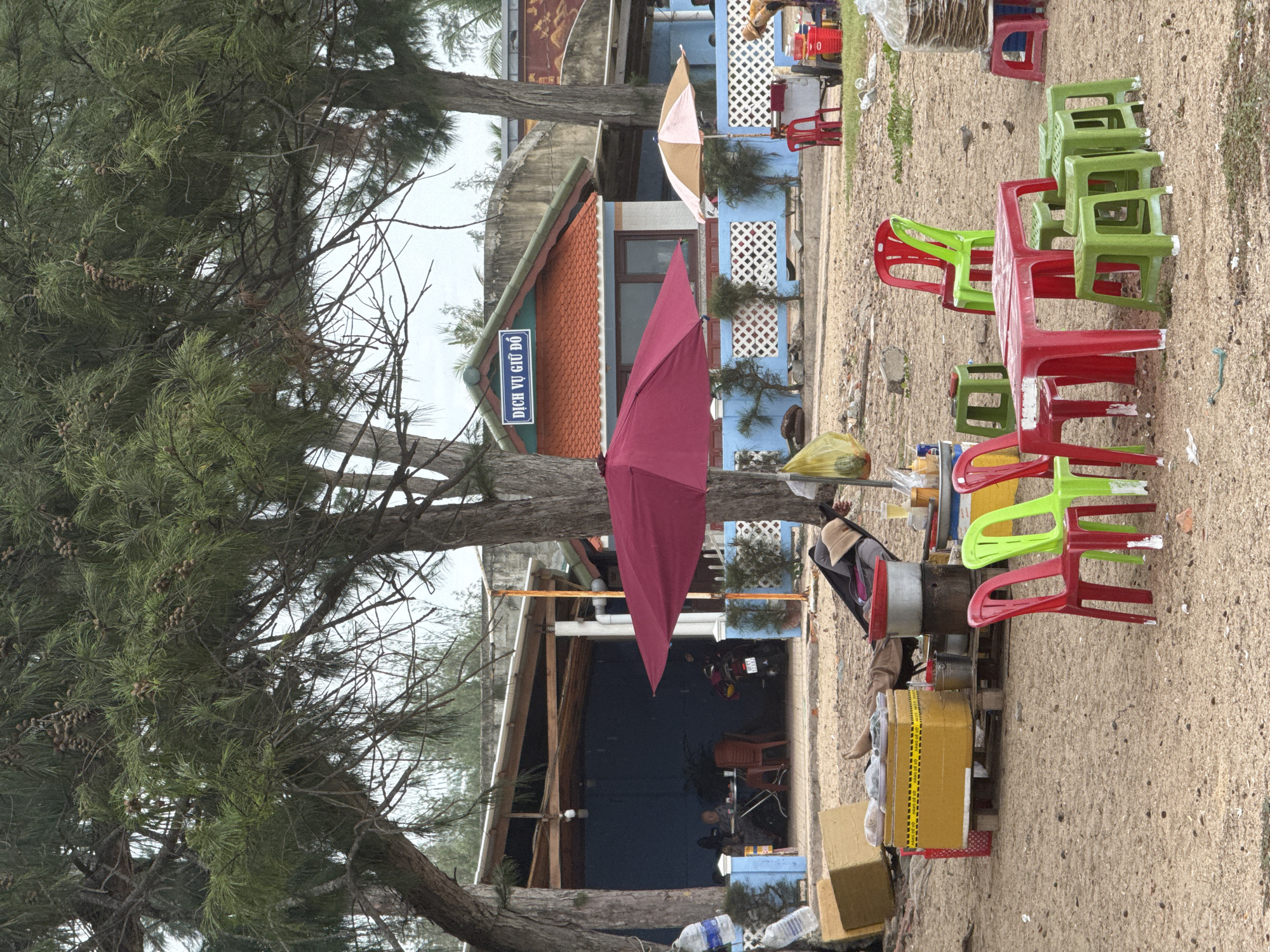 A beachside scene in Phan Thiết, Vietnam, features a collection of plastic tables and chairs, an umbrella, and food stalls set up on the sand. In the background, a building with blue walls and an orange tiled roof displays a sign reading "DỊCH VỤ GIỮ ĐỔ". Several pine trees provide shade over the area. In the foreground, a person lies in a hammock, while another person sits at a table inside the building. A motorcycle is parked near the building. The overall atmosphere is casual and rustic, typical of a local eatery or rest stop near the coast.