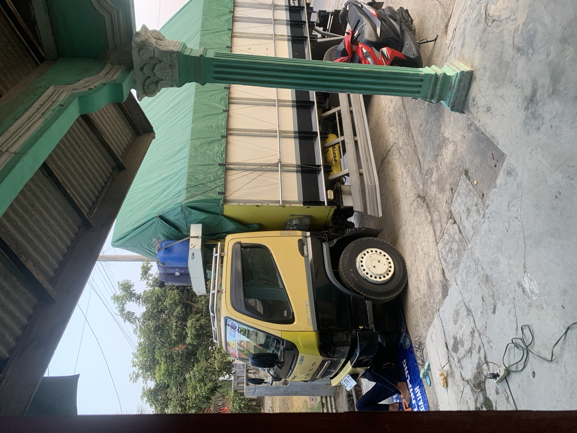 The image depicts an outdoor scene in an urban or semi-urban environment in Indonesia, likely on a street or in a yard. A large yellow and black truck is parked, its cargo area covered with a green tarp. The truck has a ladder rack on its roof, on which two large blue water containers are secured. A red motorcycle is parked next to the truck. In the foreground, a person is seen lying on a blue mat near the front of the truck, with their legs extended. An electrical extension cord is plugged into a wall socket and stretches across the concrete ground, connected to a device not fully visible in the frame. The environment includes a building with a corrugated metal roof and a decorative green pillar. Trees and some vegetation are visible in the background, along with overhead power lines. The ground is paved with concrete, showing some cracks and discoloration. The overall impression is one of a working or residential area, possibly during a daytime.