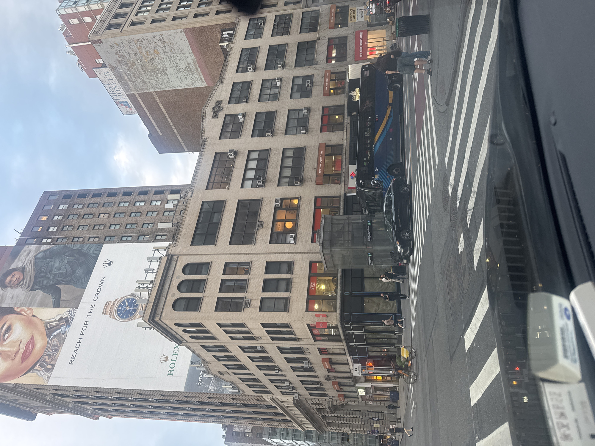 The image captures a street scene in New York City, United States of America. In the foreground, a car's dashboard is visible, suggesting the photo was taken from inside a vehicle. The middle ground shows a wide street with a crosswalk, traffic lights, and several vehicles, including a blue bus with "I ❤️ NY" branding, a black car, and a cyclist with a yellow basket. Pedestrians are also present, walking on the sidewalk and crossing the street. Buildings line the street, featuring signage for businesses such as "NYSC" (New York Sports Clubs) and "LOFT." A prominent billboard on a tall building displays an advertisement for Rolex watches and features images of Zendaya and Timothée Chalamet, seemingly related to the movie "Dune." The sky is overcast, indicating a cloudy day. The architectural style of the buildings suggests an urban environment with a mix of older and more modern structures. The overall impression is a bustling city intersection.