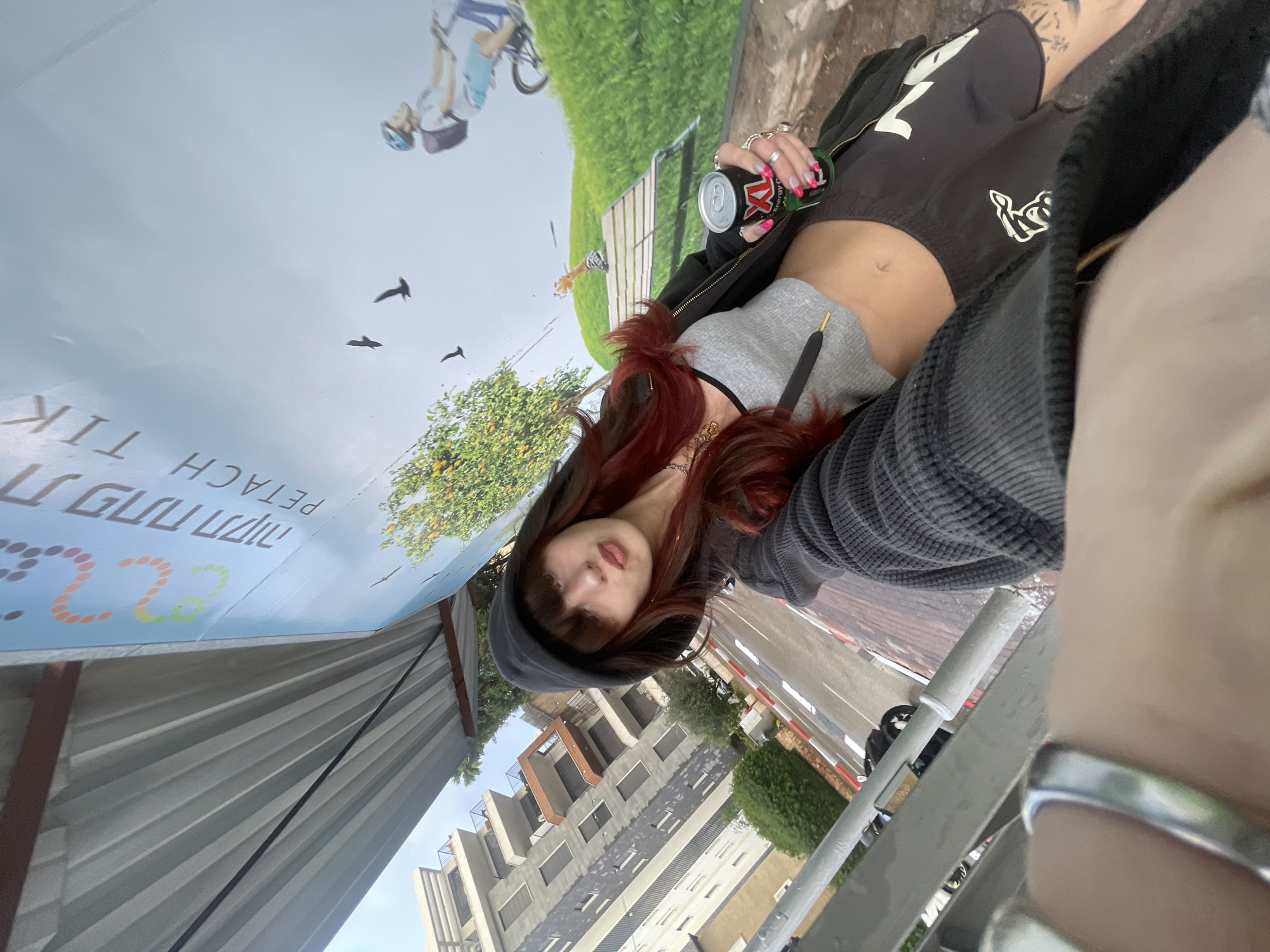 A female-presenting individual with long, reddish-brown hair and bangs, wearing a dark hooded jacket over a grey crop top and dark shorts, is depicted from the chest up taking a selfie. Her navel is visible. Her left hand holds a black and green "XL Energy Drink" can, adorned with multiple rings and bright pink painted fingernails. A small tattoo is visible on her lower left leg. The immediate background is a large, vertical sign or mural featuring green hills, orange trees, three black bird silhouettes, and a person on a bicycle, with prominent text in Hebrew "פתח תקווה" (Petach Tikva) and English "PETACH TIKVA", identifying the location in Petah Tikva, Israel. Above this sign, a corrugated metal roof structure with support beams is visible. To the subject's left, a paved street with red and white curb markings and a landscaped median containing bushes is present. Across the street, a multi-story residential building with balconies and a mixed grey and light-colored facade is visible. The subject's right hand, holding the camera, shows a silver ring on the index finger.