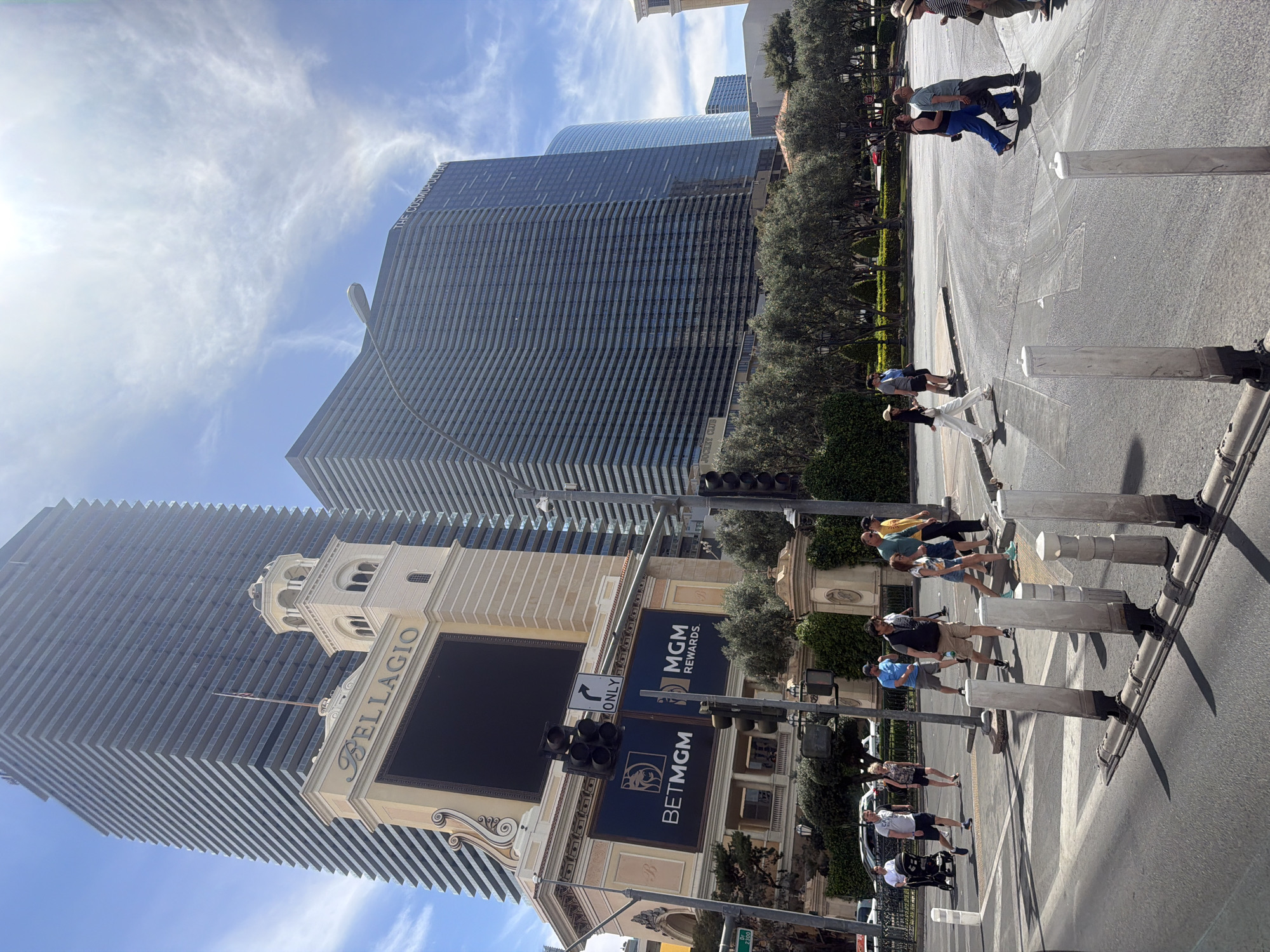 The image shows a street scene in Paradise, Nevada, United States of America, with the Bellagio hotel and casino prominently featured in the background. The Bellagio's signage, tower, and architectural details are visible, as well as the Cosmopolitan tower. People are observed crossing the street at a marked crosswalk. Traffic signals are present, and the surrounding environment includes trees and landscaping. The sky is blue with some clouds, and the sun is shining.