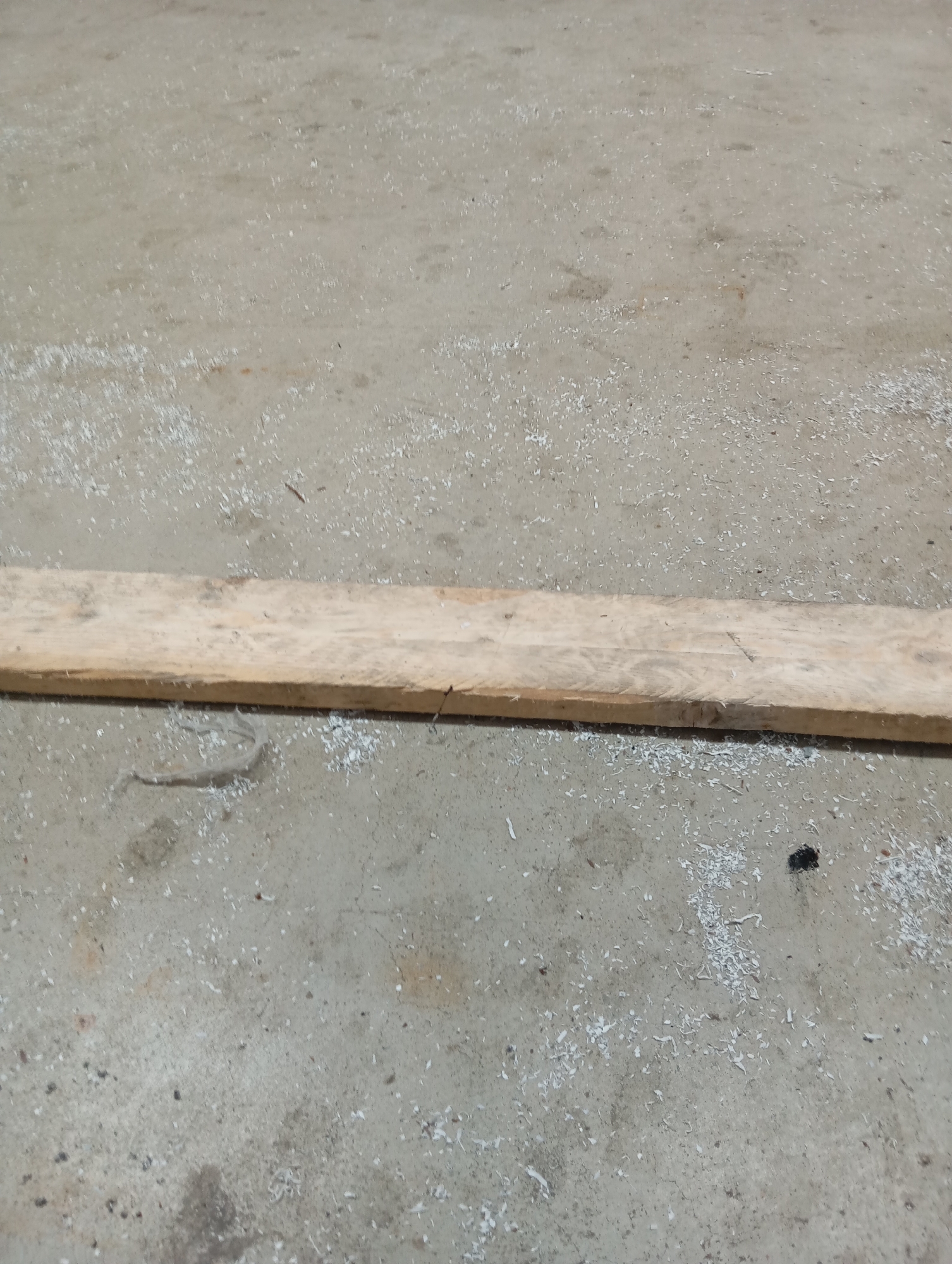 The image depicts a wooden plank lying horizontally on a concrete surface. The concrete is scattered with small white debris, possibly sawdust or wood shavings, and there are a few darker smudges and spots. The plank itself appears to be unfinished, with visible grain and some rough edges. The lighting suggests it is daytime. The scene is located in Навадворскі сельскі Савет, Belarus.