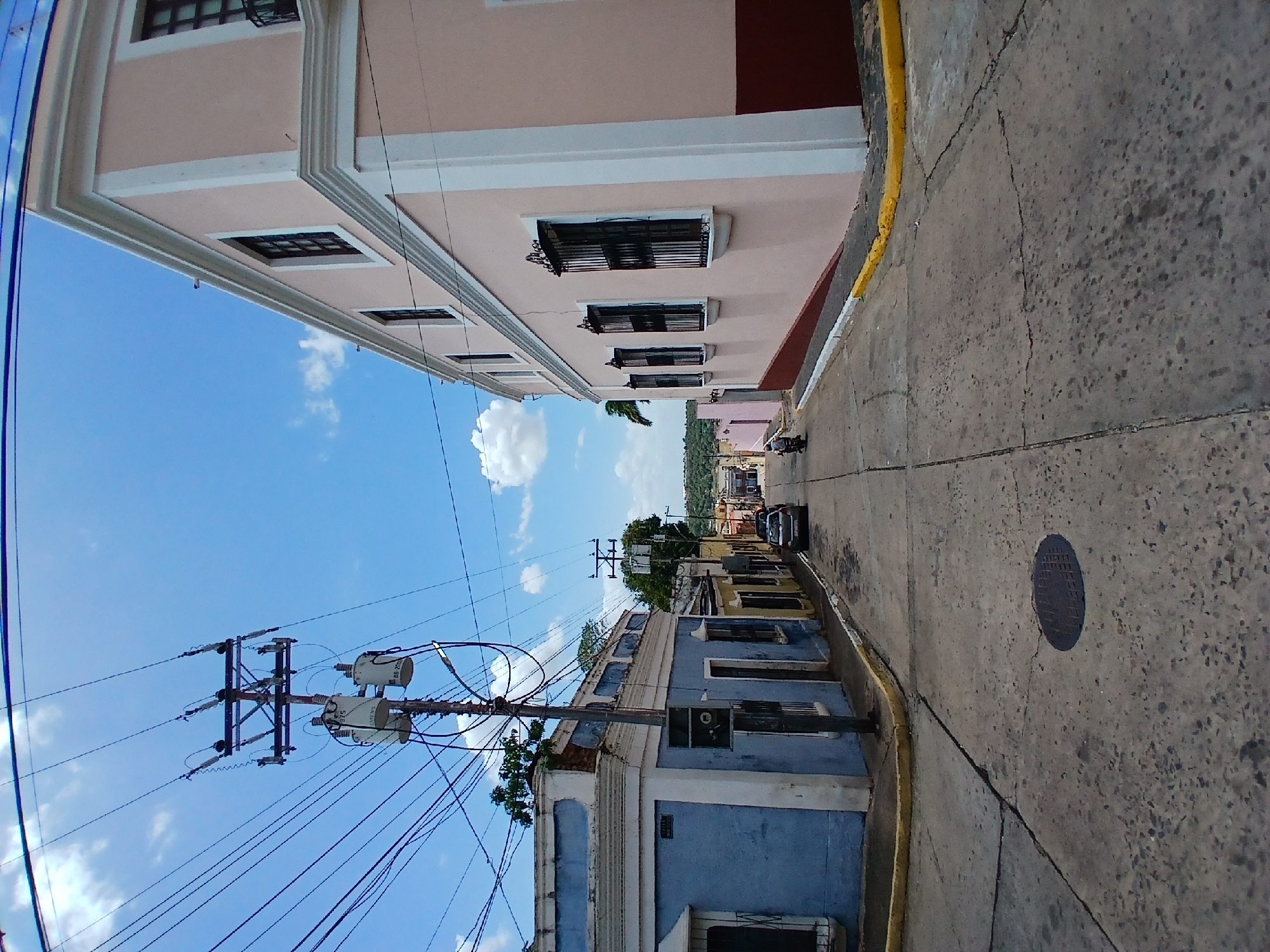 The image depicts a narrow street in Ciudad Bolivar, Venezuela, with buildings lining both sides. The building on the left side of the frame is a light pink color with a brown accent, featuring multiple windows with black bars. A yellow curb separates the building from the gray concrete road. On the right side of the frame, a blue building with white trim is visible, with utility poles and numerous wires overhead against a blue sky with scattered clouds. A manhole cover is visible on the right side of the road. A car is parked further down the street.