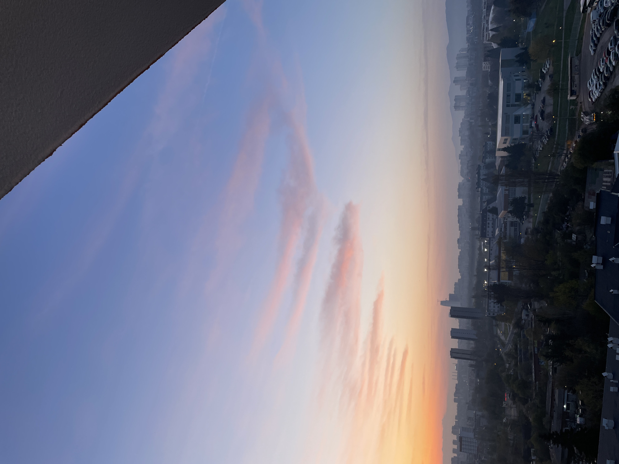 A vertical shot captures a panoramic view of the city of Sofia, Bulgaria, at either sunrise or sunset, seen from a high vantage point. The sky dominates the upper portion of the image, displaying a striking gradient of colors. It transitions from a deep, clear blue at the top, through lighter blues and purples, into warm orange, yellow, and pink hues towards the bottom-right, characteristic of twilight. Streaks of thin, elongated, reddish-pink clouds are illuminated by the low sun, stretching across the middle of the sky. A faint, white airplane contrail is visible in the upper blue section.
The top-left corner features a dark, textured, angular surface, which is likely part of a building or balcony edge from where the photograph was taken. Below the vibrant sky lies a sprawling urban landscape, partially obscured by a soft atmospheric haze or mist, which lends a muted quality to the distant features. Distant mountains are faintly visible on the far horizon, silhouetted against the colorful sky.
The cityscape includes a diverse mix of buildings, from multi-story residential blocks to commercial or industrial structures, with visible rooftops and windows. A large parking lot filled with numerous cars is discernible towards the right side of the urban area. Green spaces and trees are interspersed among the buildings. No specific activity or people are visible, and there is no visible text. The overall scene conveys a serene and expansive view of the city during the golden hour.