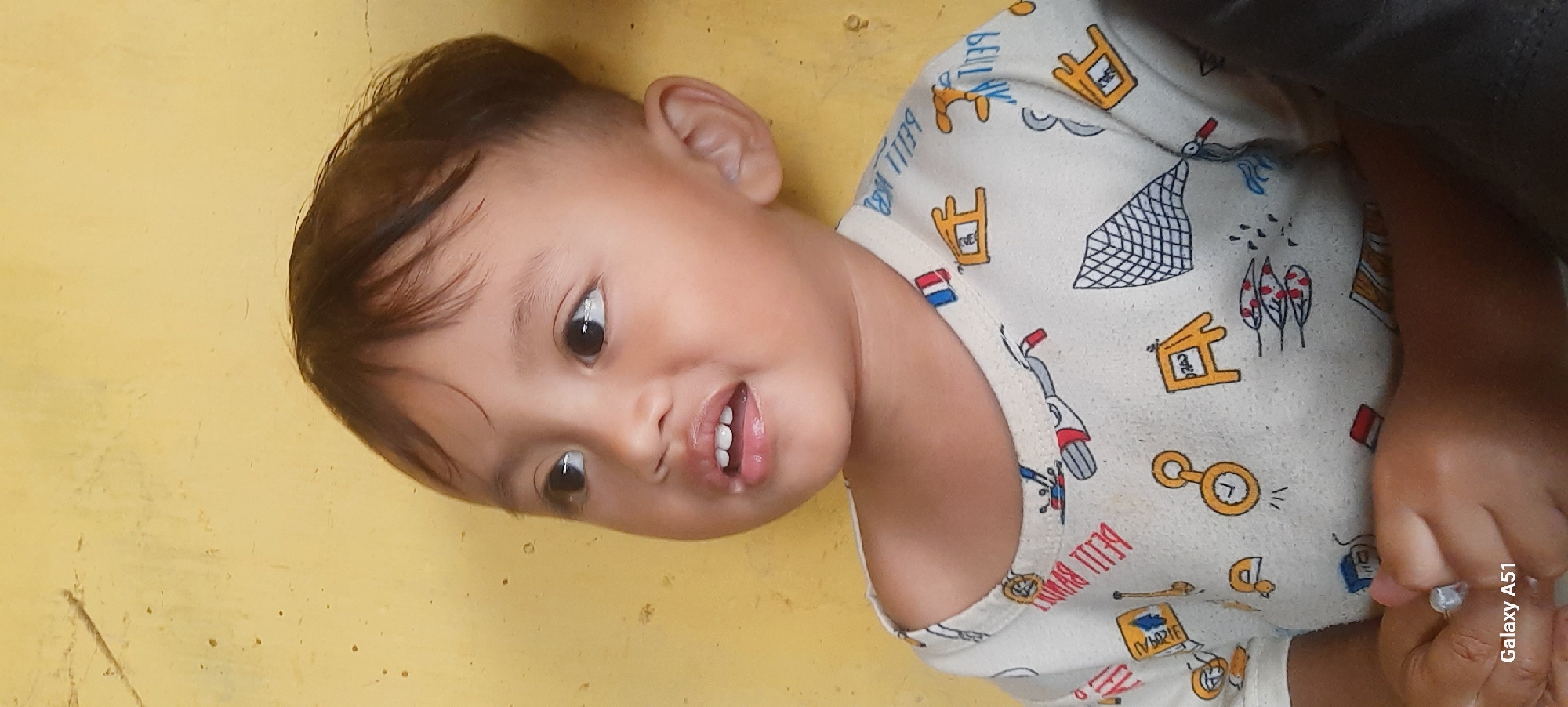 This is a close-up, sideways shot of a young child lying down. The child is looking towards the camera with a slight smile, revealing their teeth. Their eyes are wide and dark, and their hair is dark brown and short. The child is wearing a light-colored t-shirt with a pattern of various cartoonish illustrations, including what appear to be houses, trees, a bicycle, and a net. The background is a solid yellow surface, which appears to be a wall or floor, possibly with some subtle texture or markings. The lighting suggests it is daytime. The image has been captured with a Samsung Galaxy A51.