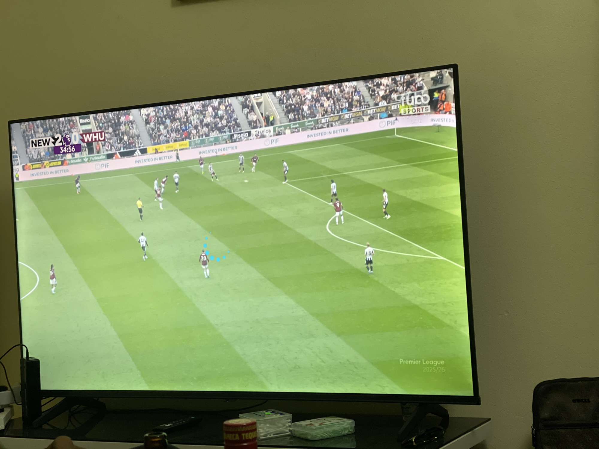 The image shows a television screen displaying a live football match between Newcastle (NEW) and West Ham United (WHU). The score is 2-0 in favor of Newcastle, and the time is 34:56. The game is being played on a green football pitch, and there are players from both teams visible on the field. The stands are filled with spectators. The bottom of the screen displays "Premier League 2025/26."
The television is sitting on a table, and there are several objects visible on the table in front of the screen, including a bottle, boxes, and headphones. Part of a bag is visible in the lower-right corner. The image does not provide information about the location (Abuja, Nigeria).