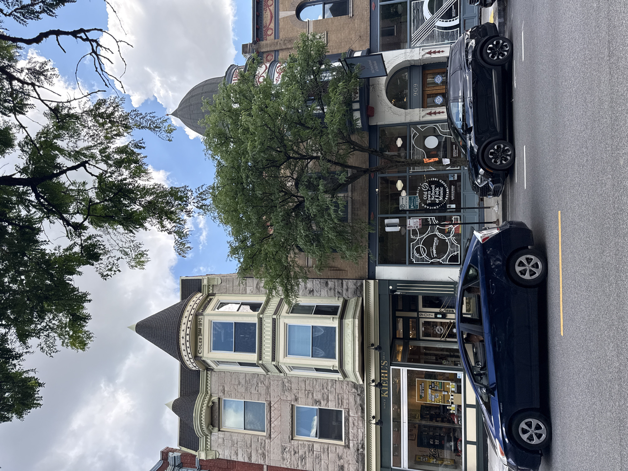 This media file depicts a street scene in Chicago, United States of America, likely during the daytime under a cloudy sky. The image is oriented vertically, with the street running from the bottom to the top of the frame. The foreground features a dark blue hybrid vehicle parked on the right side of the street, parallel to the curb.
The buildings lining the street are a mix of architectural styles. On the left, a historic building with stone facade and a conical turret is visible, with the text "COLES B-1890." inscribed on it. Further up the street, a building with a dome and ornate details can be seen, featuring signage for "Old Town School of Folk Music" and "ESTA 1957". The building next to it has a sign indicating the address "909". Another building further up displays the number "907". On the right side of the street, a black SUV is parked.
A large tree with lush green foliage is prominently positioned in the center of the image, partially obscuring the buildings and the sky. The presence of trees and the architectural styles suggest an urban environment with a historical character. The sky is partly cloudy, indicating moderate weather conditions. There are no people clearly visible in the frame, and the overall mood is quiet and observational, focusing on the architecture and the streetscape. Visible text on the buildings and signs provides specific details about the establishments and their history.