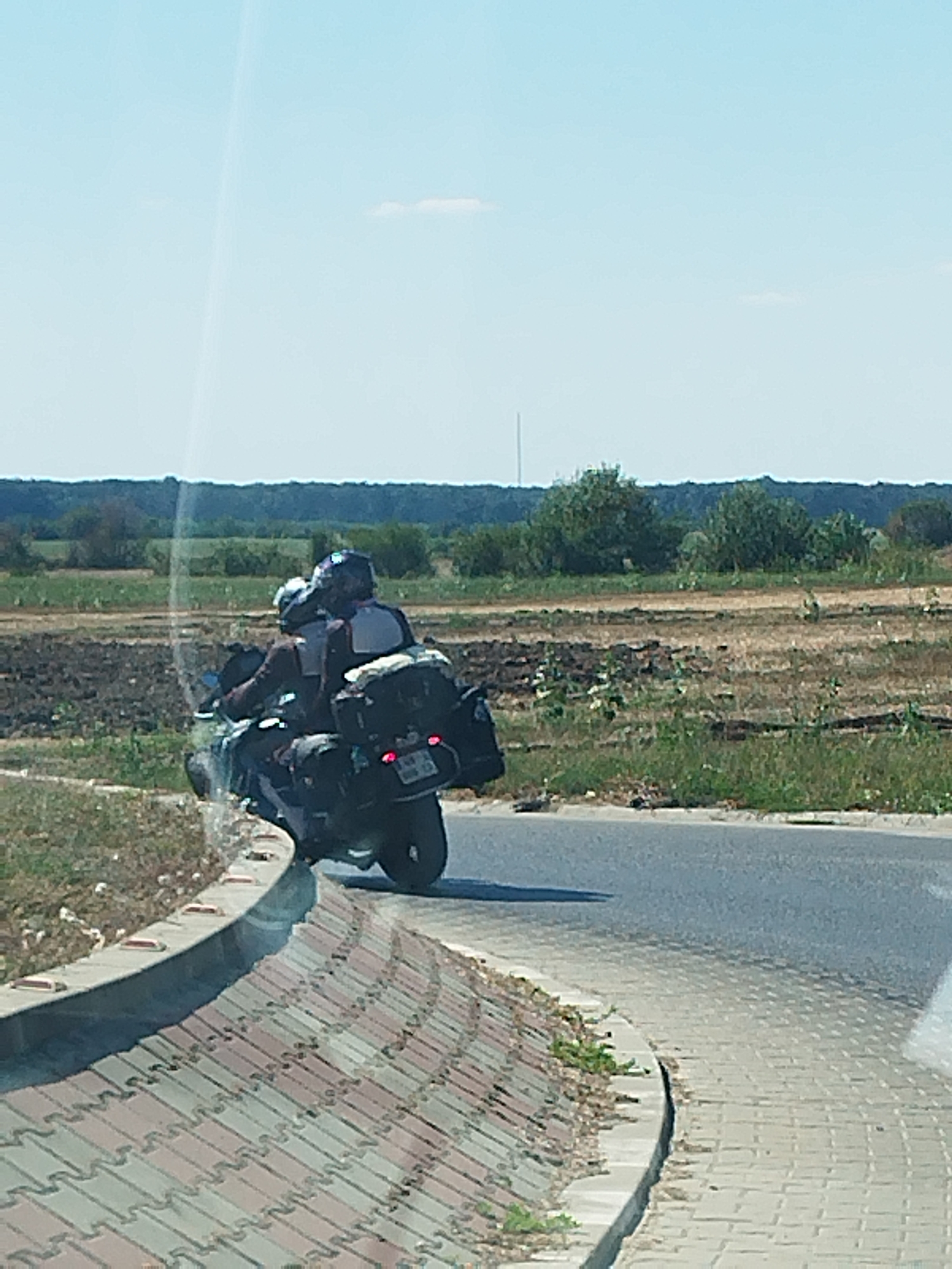 The image captures a scene on a road in Buftea, Romania, under a clear sky. Two individuals, wearing helmets and protective gear, are riding a motorcycle equipped with saddlebags. The motorcycle is in motion, leaning into a curve on the right side of the paved road. The road is lined with a decorative brick wall on the left, which curves with the road. Beyond the wall and road, there are fields with patches of dry earth and vegetation, leading to a line of trees in the distance. The overall impression is a snapshot of a motorcycle journey on a sunny day in a rural or semi-rural setting.
