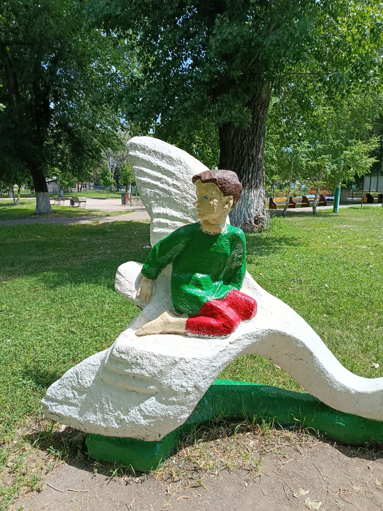 A concrete statue of a young boy with brown hair, blue eyes, wearing a green shirt and red pants sits on a white, abstract, dragon-like form. The sculpture is located in a park area in Marx, Russia, with a grassy lawn, trees, and playground equipment visible in the background. The statue is painted in bright colors, and the dragon form has a green base. The lighting suggests it is daytime, likely late morning or early afternoon, with dappled sunlight filtering through the trees.