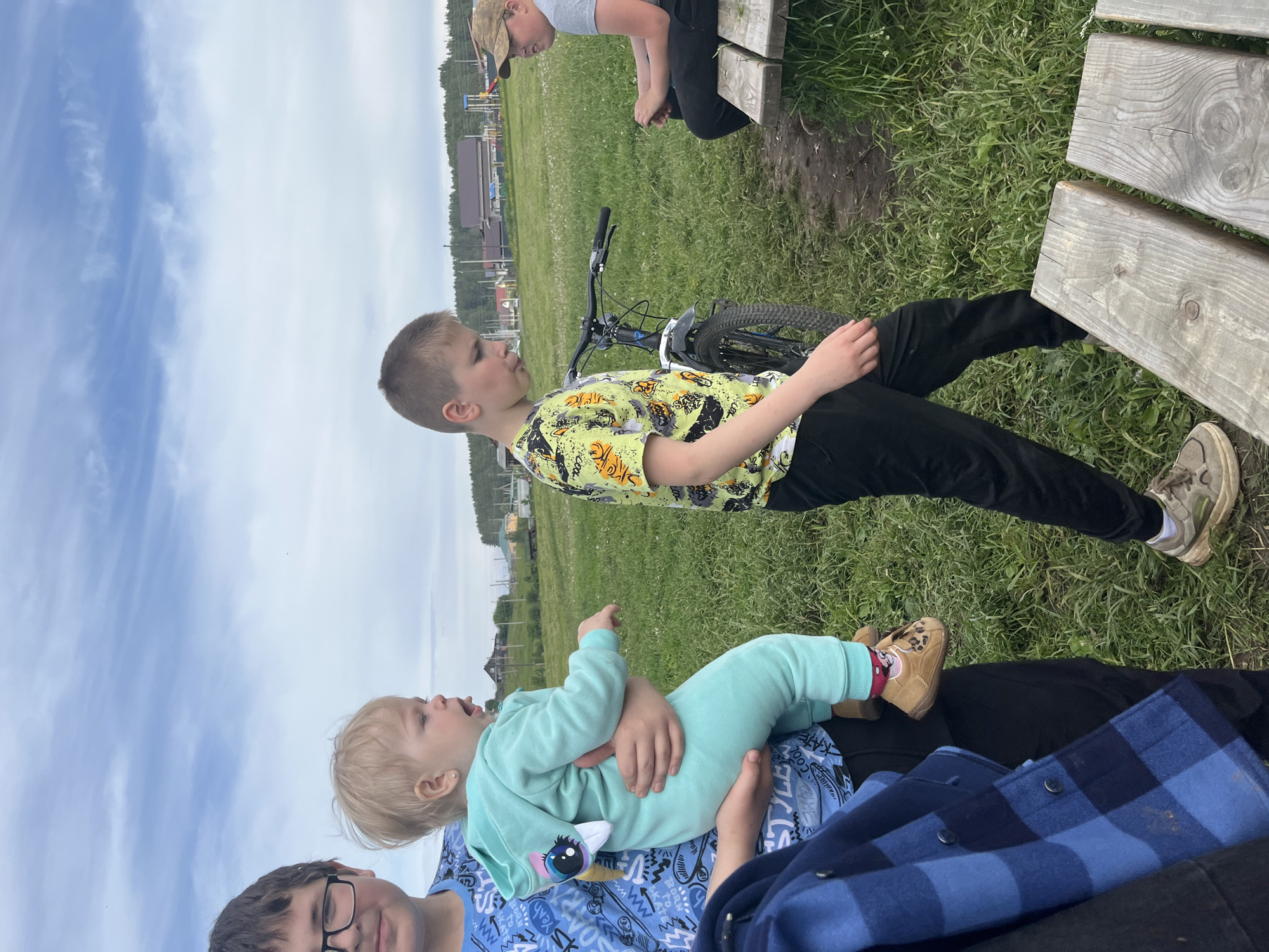 This image, taken in Фоки, Russia, depicts an outdoor scene featuring several individuals in a grassy area under a partly cloudy sky during the daytime.
In the lower-left foreground, a person with dark hair and glasses, wearing a blue patterned shirt and a blue plaid overshirt, is holding a blonde-haired toddler. The toddler is dressed in a light blue jumpsuit featuring a unicorn design on the chest, along with brown shoes and red socks. The toddler is looking upwards with an open mouth, suggesting they might be speaking, singing, or reacting to something.
To the right, a young boy with short brown hair, wearing a yellow-green t-shirt adorned with animal patterns (possibly tigers) and black pants, stands beside a black mountain bicycle. He is looking to his left.
Further in the background, towards the upper right, another individual (likely an older boy or teenager) wearing a cap, a grey t-shirt, and black pants, is seated on a rustic wooden bench. Their hands are clasped together.
The setting is a green field with some wild grasses and a small area of exposed dirt near the bench. In the distance, structures resembling houses or village buildings are visible, interspersed with trees, consistent with a rural or semi-rural landscape. The overall mood appears casual and relaxed, indicative of an outdoor gathering or leisure activity. Some indistinct patterns are visible on clothing, but no clear text is legible.