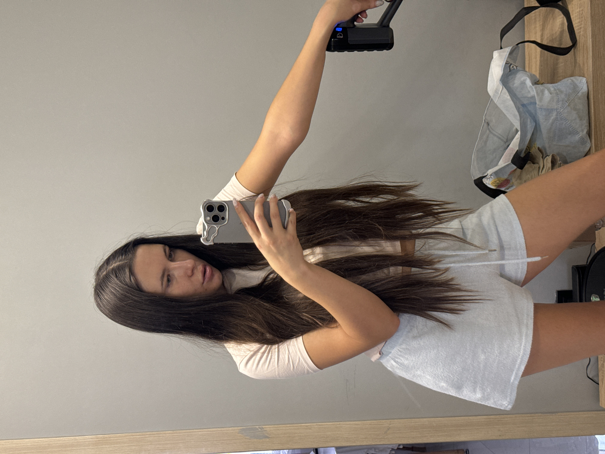 A young woman is captured in a mirror selfie, holding a smartphone in her left hand to take the picture. Her long, dark brown hair flows over her shoulders and chest. She is wearing a light-colored, short-sleeved top and light grey or white drawstring shorts. Her expression is neutral as she looks towards the phone.
In her raised right hand, she holds a black electronic device with a handle, possibly a portable light or camera accessory, which exhibits blue indicator lights. To the right of the frame, a light-colored fabric tote bag with black straps hangs, containing some items. The background is a plain light grey wall, and below the mirror, a light-colored wooden surface is visible. The scene is indoors, in what appears to be a room within a residence in Odintsovo, Russia. No discernible text is visible.