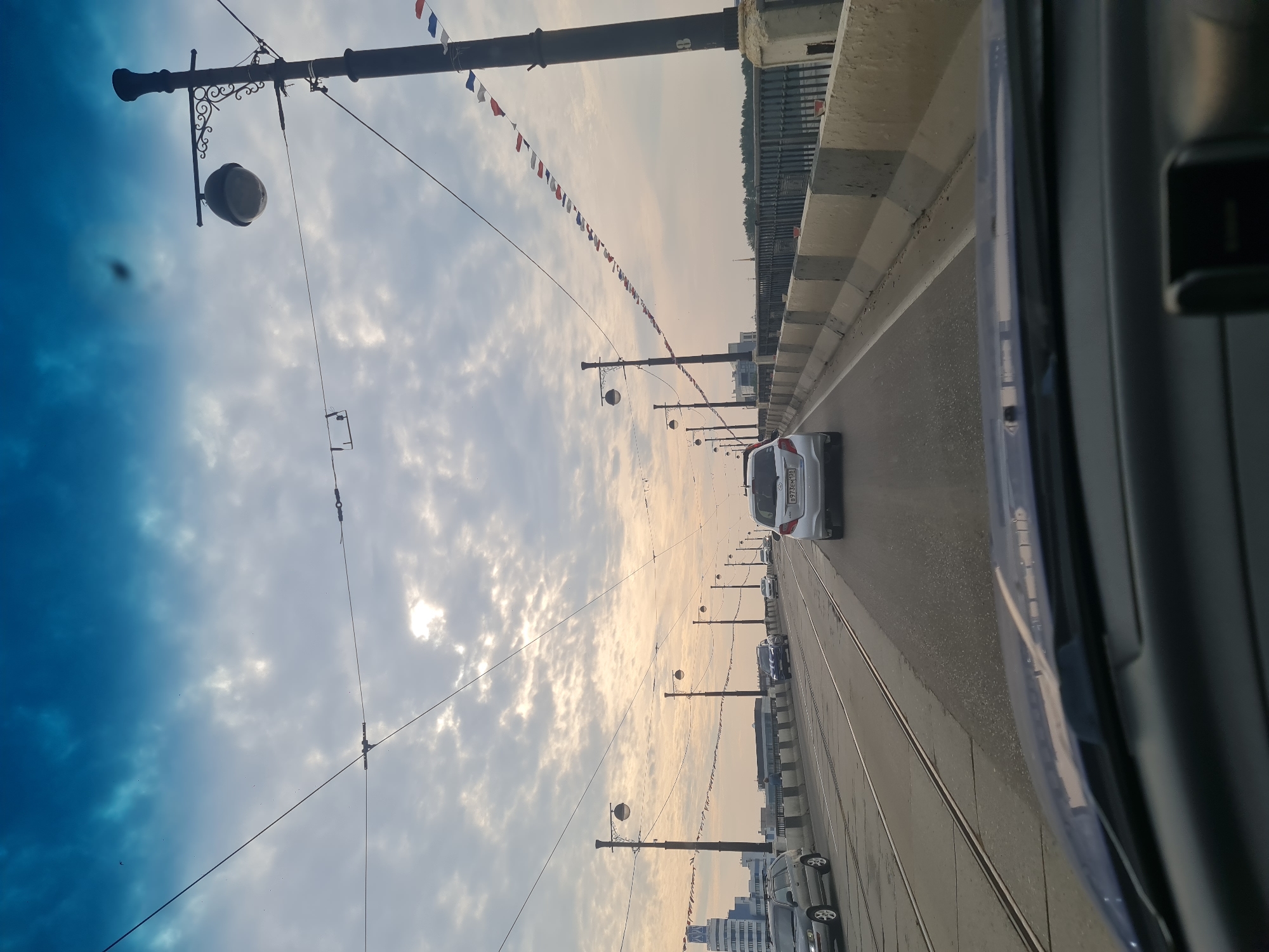 A photograph taken from the interior of a vehicle, looking forward through the windshield, captures a view of a bridge on a cloudy day. The bridge has multiple lanes of traffic and tram tracks. Several cars are visible, including a silver sedan in the foreground, a silver SUV on the left, and other vehicles further ahead. The bridge is lined with decorative lampposts and a railing. A string of small red, white, and blue flags hangs across the sky, attached to wires overhead. In the distance, a tall, modern building with a blue facade is visible on the left side of the frame. The sky is a mix of blue and gray clouds, with sunlight breaking through in places. The image was taken on the Alexander Nevsky Bridge in Irkutsk, Russia.