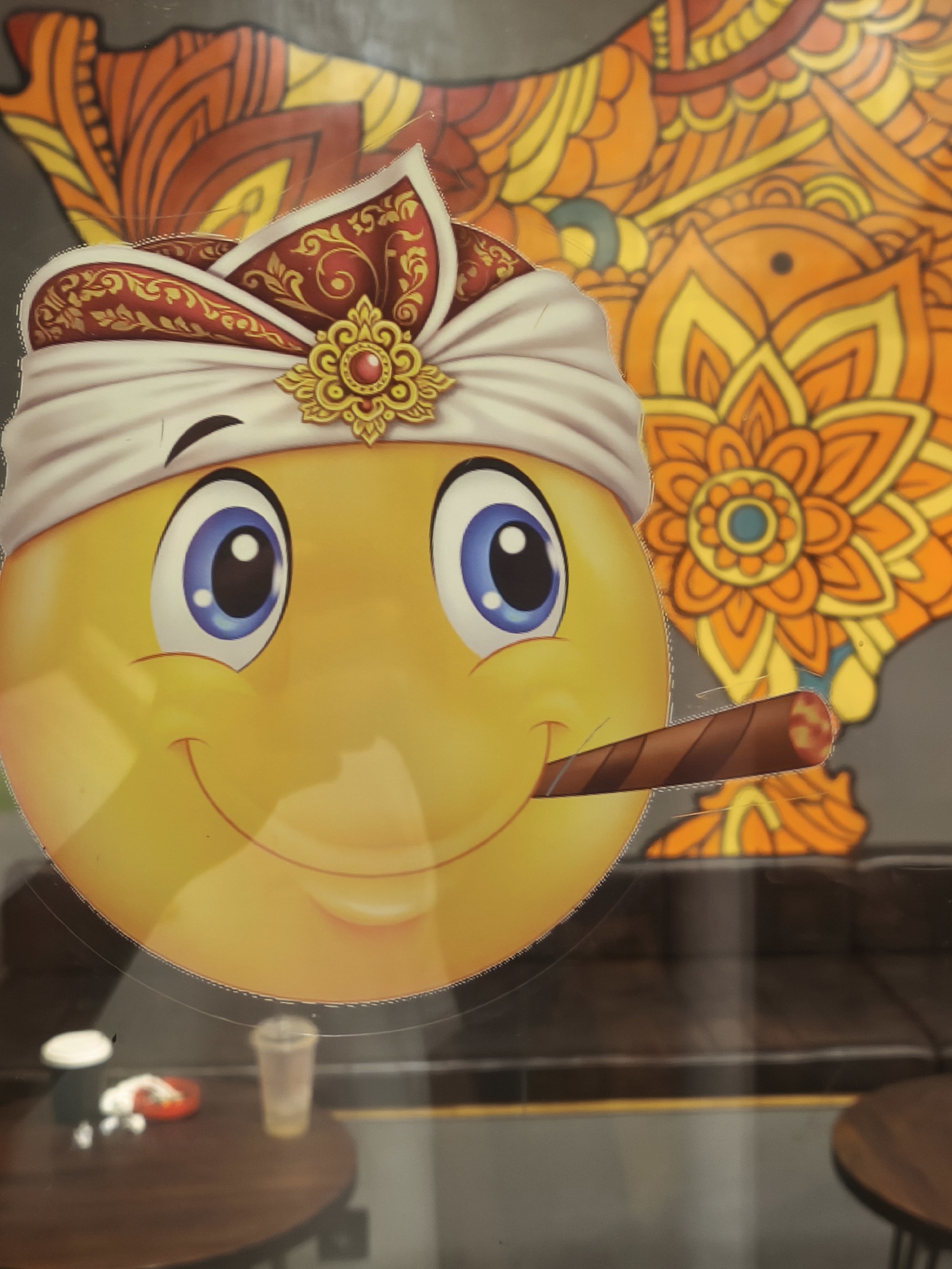 This is a medium-shot photograph of a large, glossy yellow emoji sticker with a cigar in its mouth, attached to a window or glass surface. The emoji wears a white and red turban with gold ornamentation and a red jewel. Behind the emoji is a colorful, stylized orange and yellow floral or paisley design on a dark background. The emoji's face is depicted with large blue eyes, a winking left eye, and a smiling mouth. The cigar is brown with red and yellow at the tip, suggesting it is lit. The lower portion of the image shows a blurry view of a dark wooden table with a few objects on it, including a small white object, a red and white object, and a clear plastic cup with some liquid. The overall impression is that this is a decorative sticker in a commercial establishment, possibly a cafe or shop, in Kuta, Indonesia. The background suggests an indoor setting with a reflection on the glass.