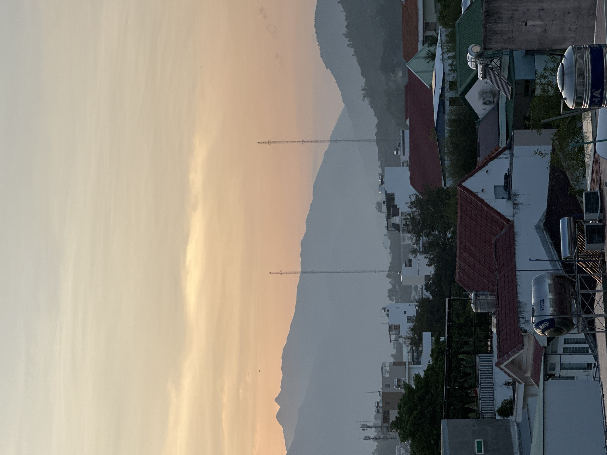 The image depicts a panoramic view of Bac Nha Trang Ward, Vietnam, at either sunrise or sunset, characterized by a gradient sky transitioning from light blue at the zenith to warm orange and yellow hues near the horizon. Wispy clouds are subtly present across the sky, with two small, dark bird silhouettes discernible in the upper mid-ground.
In the mid-ground, a prominent range of mountains stretches across the horizon, appearing in muted blue-grey tones due to atmospheric perspective. The mountains on the left feature distinct, jagged peaks, while those on the right are smoother. Two tall, slender lattice communication or transmission towers stand silhouetted against the sky, one positioned to the left of the central mountain range and another further right.
The foreground and lower mid-ground reveal a dense residential area. Numerous buildings, predominantly houses, feature a mix of pitched roofs in terracotta red and dark green, alongside flat concrete rooftops. On these rooftops, various utility installations are visible: multiple large, cylindrical or spherical water tanks made of metallic (stainless steel) material, some bearing visible branding such as "SONHA," "MX," and "1000L" capacity markings. Several solar water heaters, consisting of a panel and a horizontal tank, are also installed on rooftops. Outdoor air conditioning compressor units are mounted on building exteriors and rooftops. Green vegetation, including trees and dense foliage, is interspersed throughout the urban landscape, suggesting a mix of residential structures and greenery. No people or overt actions are visible in the scene.