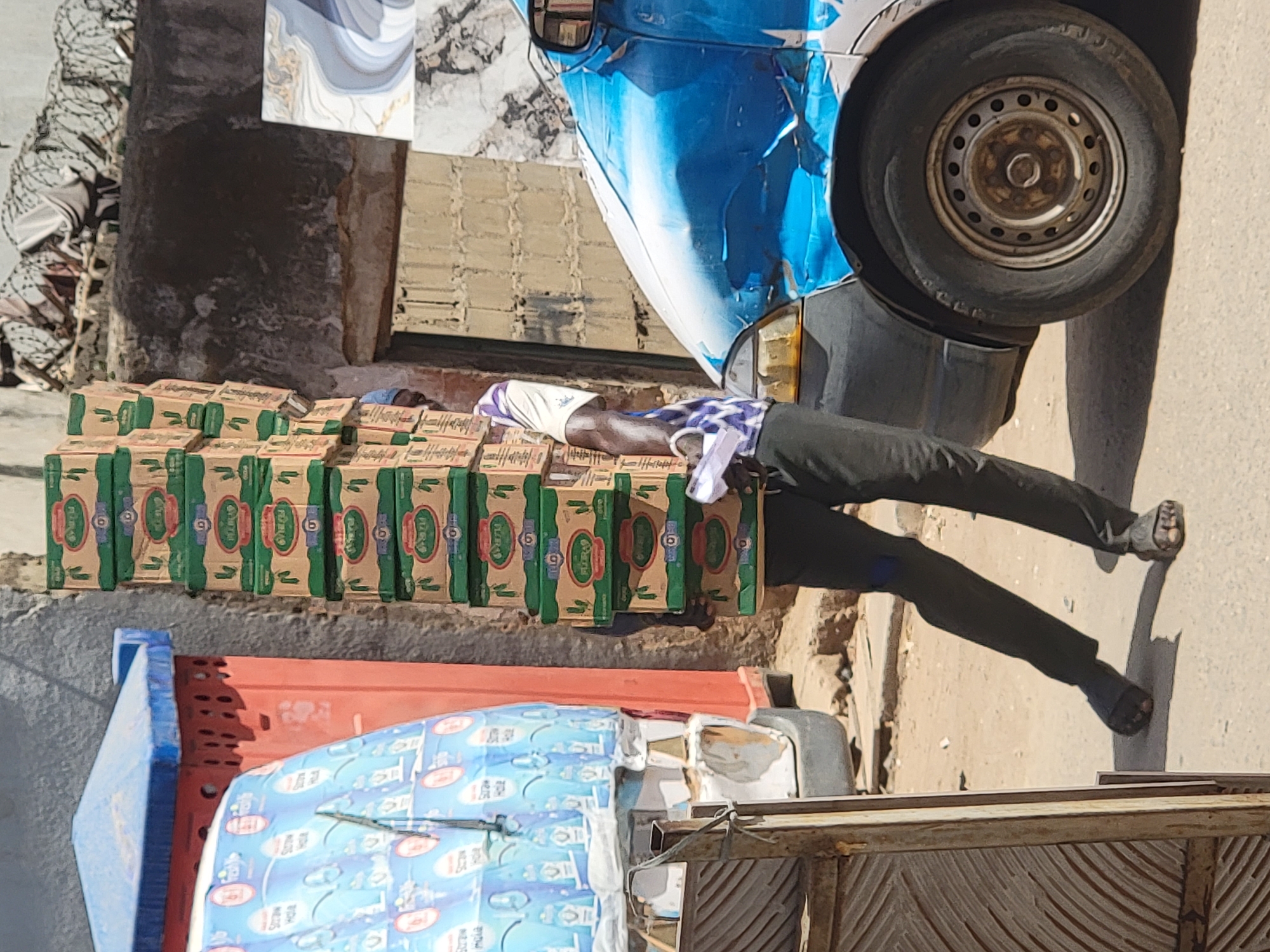 This vertical full shot captures a street scene, likely in Maiburiji, Nigeria, under bright daylight with strong shadows indicating a sunny day, possibly midday or early afternoon.
The central subject is a person lying on stacks of cardboard boxes. The individual is wearing dark trousers, a patterned blue and white shirt, and a white head covering. They appear to be resting or sleeping, with their body stretched out over multiple rows of boxes. Their feet, clad in simple sandals or barefoot, extend towards the right side of the frame.
Adjacent to the resting person is the front section of a light blue and white van, which shows signs of wear, possibly peeling paint or minor damage. One of its front wheels is visible on the right.
The boxes the person is resting on are primarily green and brown, with the brand name "FLORA" and the number "6" clearly visible on their sides. These boxes are neatly stacked in several rows, forming a makeshift bed. In the foreground, at the bottom left, there is another structure, possibly a cart or another vehicle, carrying items covered in a light blue patterned material. Text visible on this material includes "BOA" and "mula," suggesting it might be related to milk or water products.
The ground is unpaved and appears dusty. In the background, there are various wall textures, including some patterned tiles and rougher concrete surfaces, suggesting an urban or semi-urban setting, possibly near a market or a street-side commercial establishment.