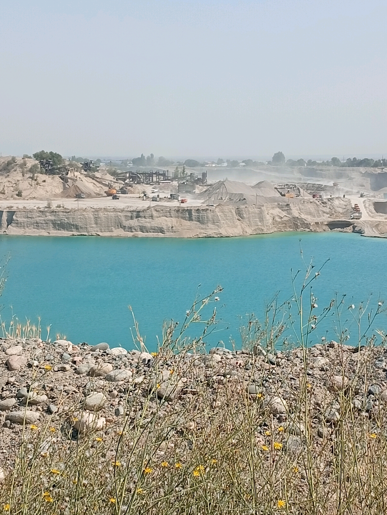 The image depicts a quarry with a body of turquoise water. In the background, industrial equipment and mounds of earth are visible, suggesting ongoing excavation or construction activities. The quarry appears to be located in or near the city of Бәйтерек, Kazakhstan, given the context. The foreground is occupied by a rocky embankment covered in sparse vegetation. The sky is hazy and light blue. There are no people visible in the image.