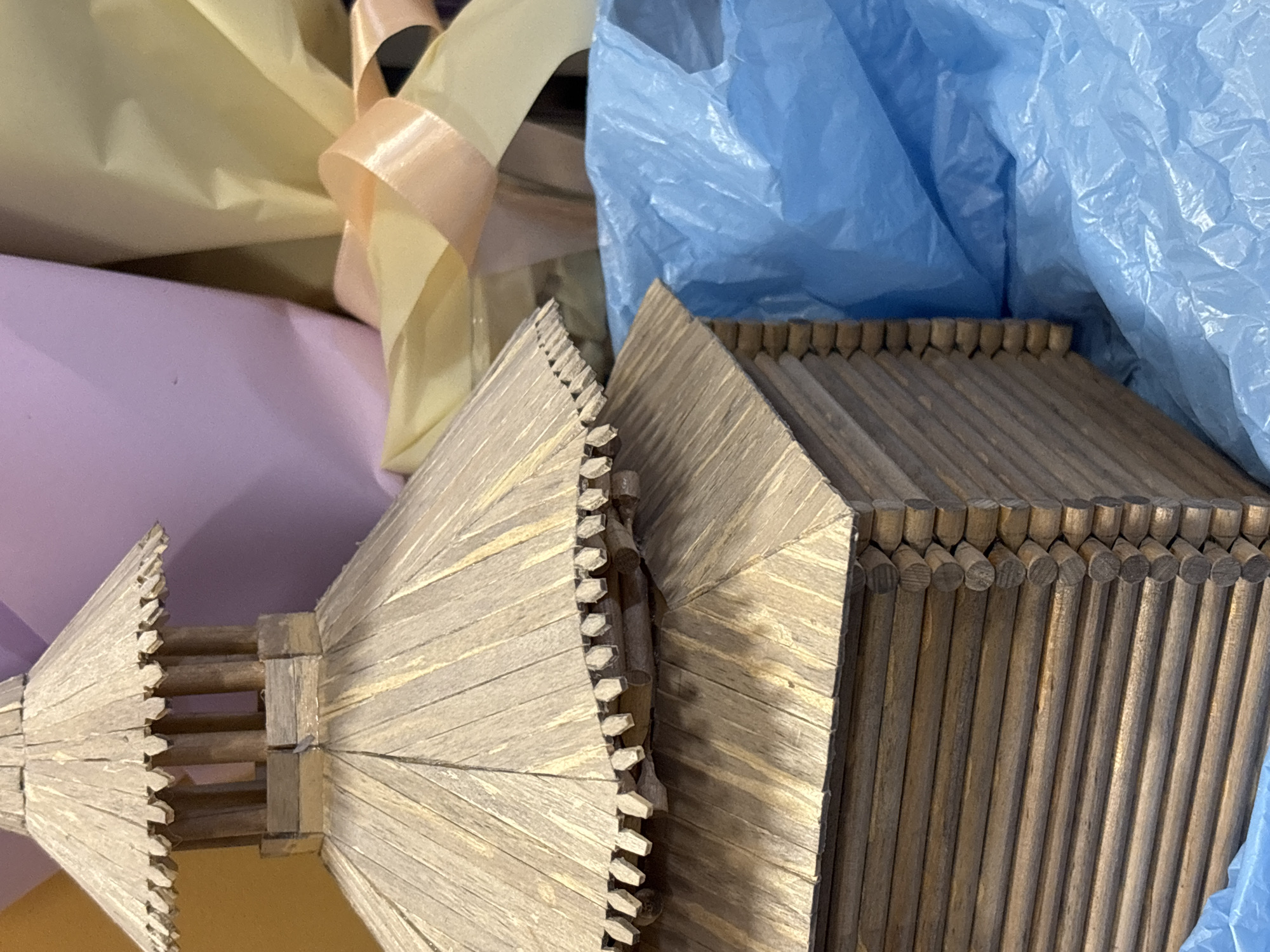 The image shows a close-up of two wooden models, likely houses or architectural structures, constructed from what appear to be small wooden sticks or dowels. The structures have a weathered, aged appearance. The models are surrounded by wrapping materials, including a light blue crinkled plastic sheet, yellow, and light pink colored papers and ribbons. Given the description as being located in Perm, Russia, these structures are possibly replicas of historical Russian architecture. The scene appears to be an indoor setting. No people are visible.