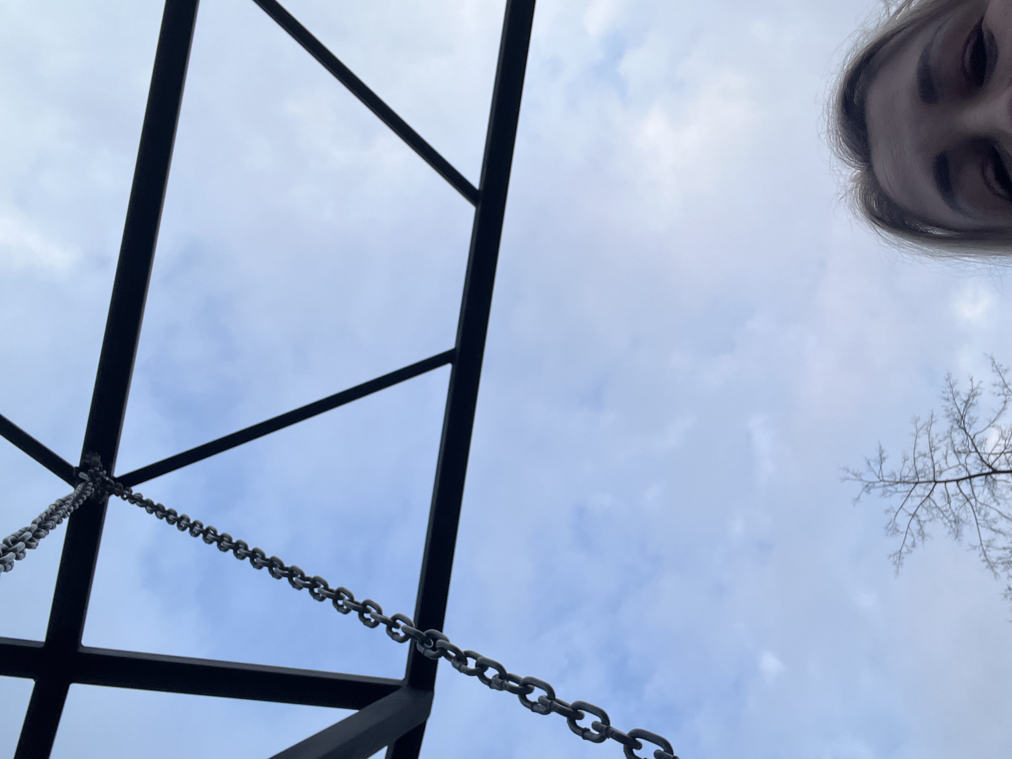 The image presents a low-angle view looking upwards. A portion of a person's face is visible in the top right corner of the frame. The majority of the image is dominated by a pale blue sky with scattered clouds. In the lower-left quadrant, a black metal structure is visible, possibly part of a swing set or playground equipment, with a chain hanging from it. Bare tree branches are seen in the lower-right, contrasting against the sky. Without additional information, it's impossible to confirm the specific location within Chaikovsky, Russia, or definitively identify any activities or interactions.