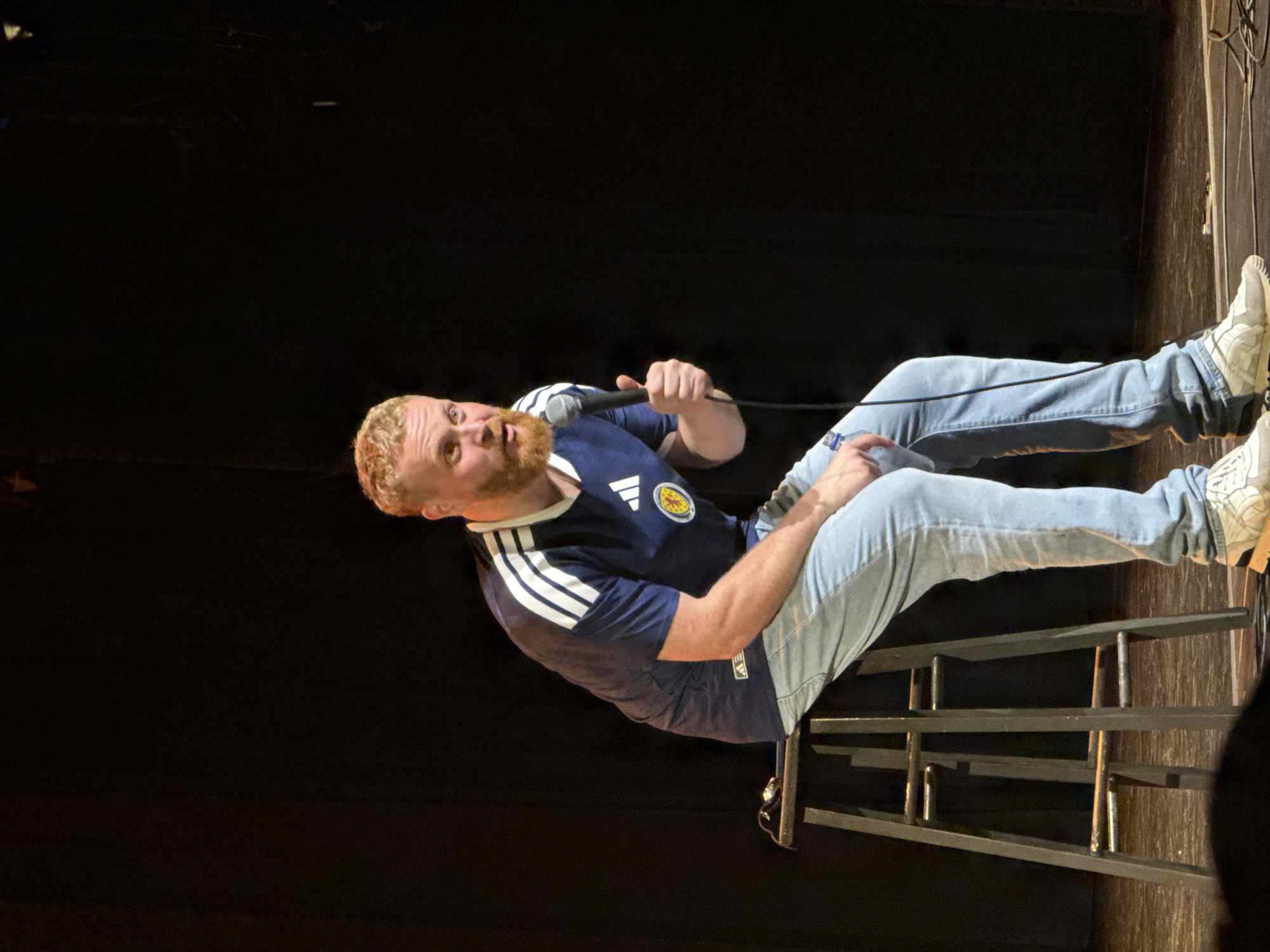 A white man with reddish-blonde hair and a beard is sitting on a stool on a stage. He is wearing a navy blue shirt with white stripes on the sleeves and a circular emblem on the left chest, and light blue jeans. He is holding a microphone in his right hand and appears to be speaking or performing. The background is dark and indistinct, suggesting a stage setting. The floor of the stage is visible and appears to be wood. There are some cables on the floor in the lower right corner of the image. The stool is a dark metal bar stool with a wooden seat. There is a small black object, possibly a phone, on the seat of the stool next to the man. The lighting appears to be from the front, illuminating the man and the stool.