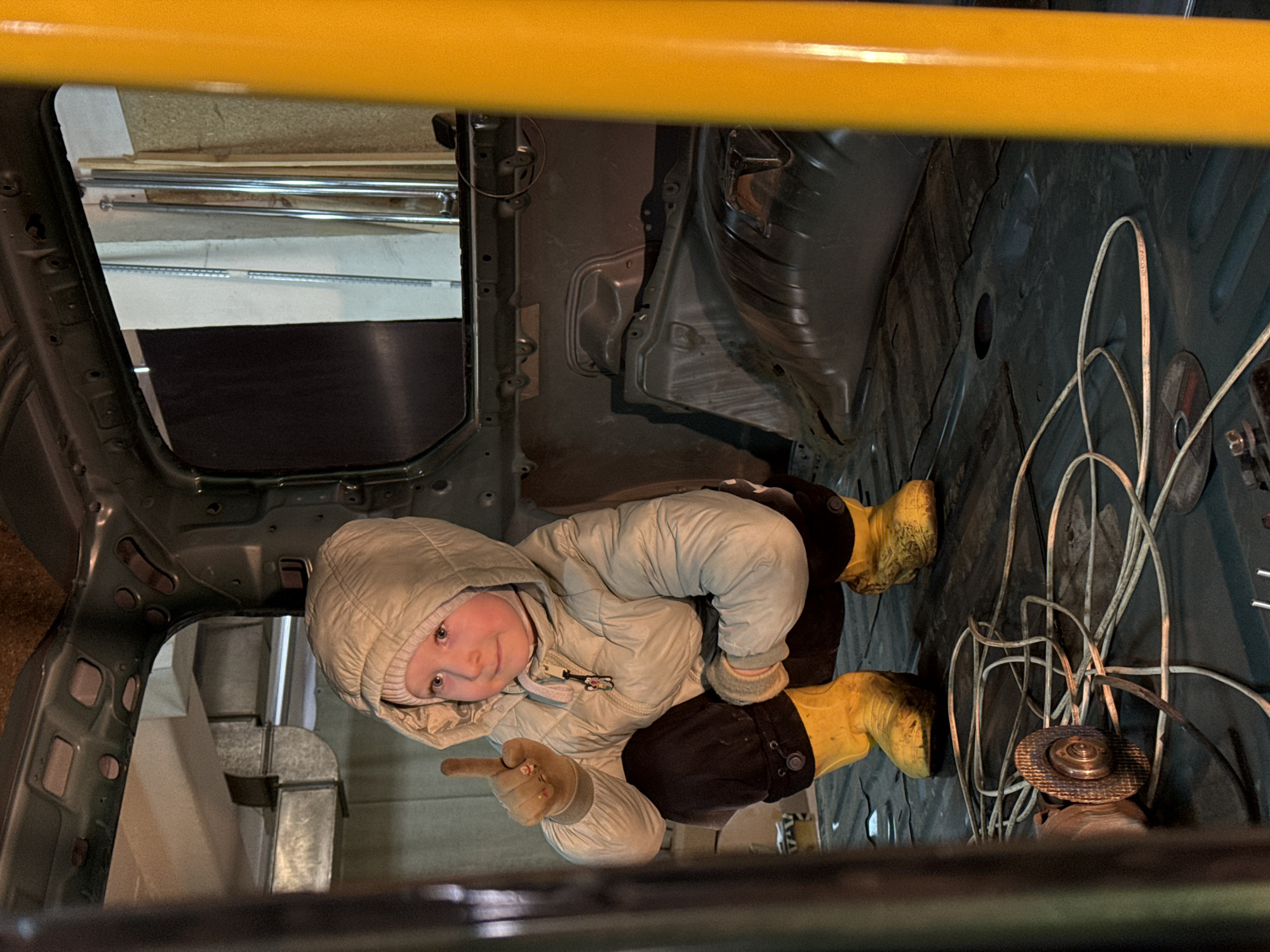 A child in a hooded jacket and yellow boots is crouched inside the stripped interior of a vehicle, possibly undergoing restoration. The location is likely a garage or workshop in Perm, Russia. The interior of the vehicle is bare, exposing metal framework and flooring. An angle grinder and wires are visible near the child. The child is looking up and pointing towards the viewer.