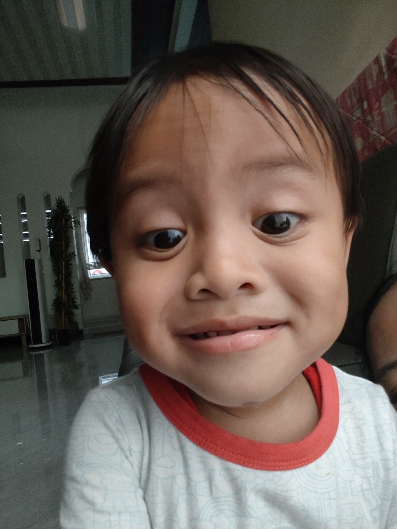 A close-up, slightly distorted, forward-facing selfie of a young child with dark hair and brown eyes. The child is smiling with lips parted and teeth visible. The child is wearing a white t-shirt with a red neckline and a light blue pattern of abstract shapes. The background appears to be an indoor setting with a tall potted plant to the left and a window with sheer curtains to the right. The flooring is reflective. There is no information in the image to determine the location in Pemalang, Indonesia.