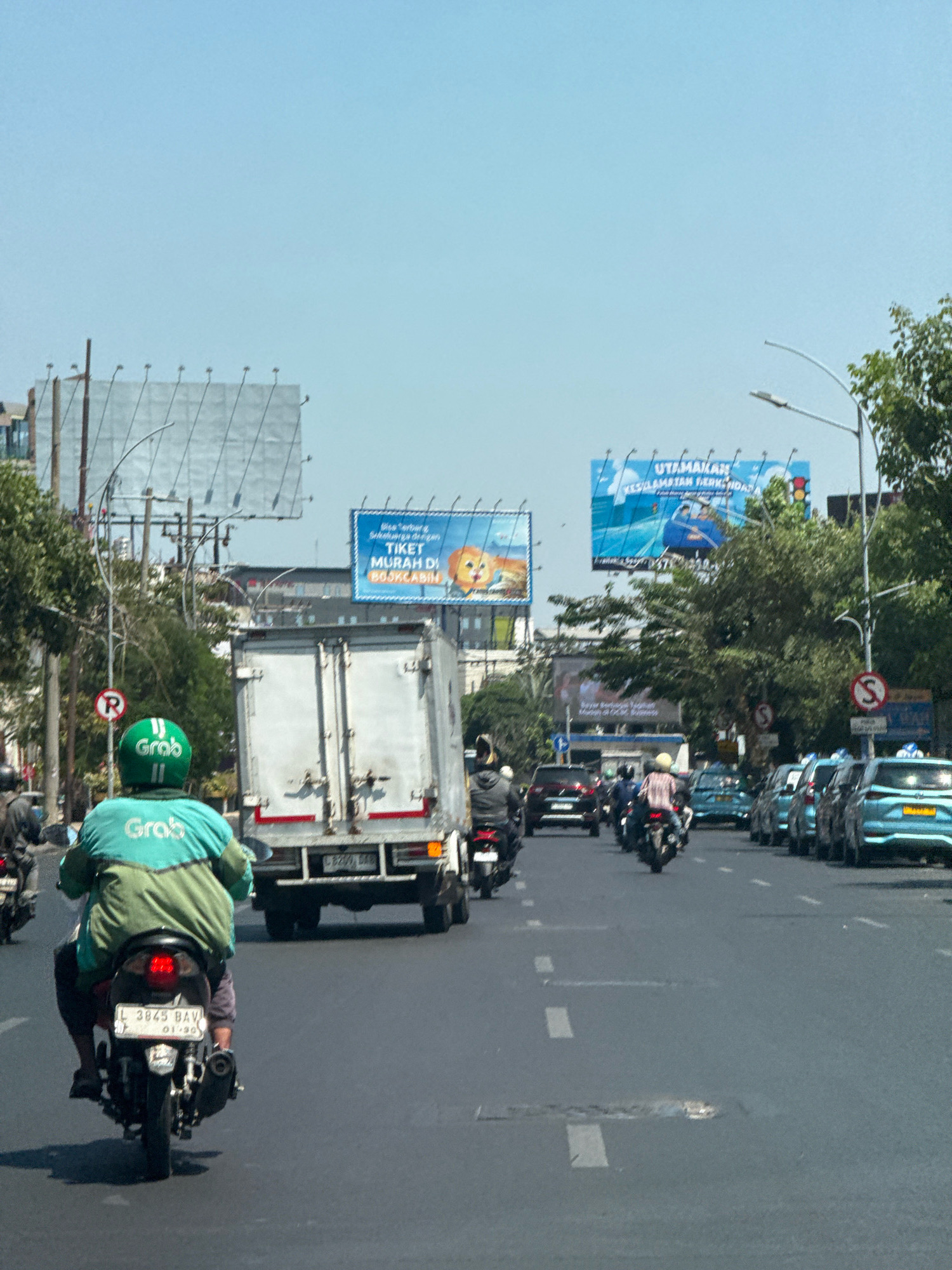A street scene in Surabaya, Indonesia, captures a sunny day with clear blue skies. The image is dominated by a road filled with various vehicles, including motorcycles, cars, and a large truck. In the foreground, a motorcycle rider wearing a green Grab jacket and helmet is moving away from the camera. The motorcycle has a license plate that reads "L 3845 BAV". A large white truck is positioned in the middle of the road, to the right of the rider. Further down the road, a line of light blue cars is visible, some with taxi markings. Several billboards are also present, with one on the right displaying an advertisement for "TICKET MURAH DI BOSKABIN". The environment appears to be urban, with trees and buildings lining the street. The overall impression is of a bustling city street during daylight hours.