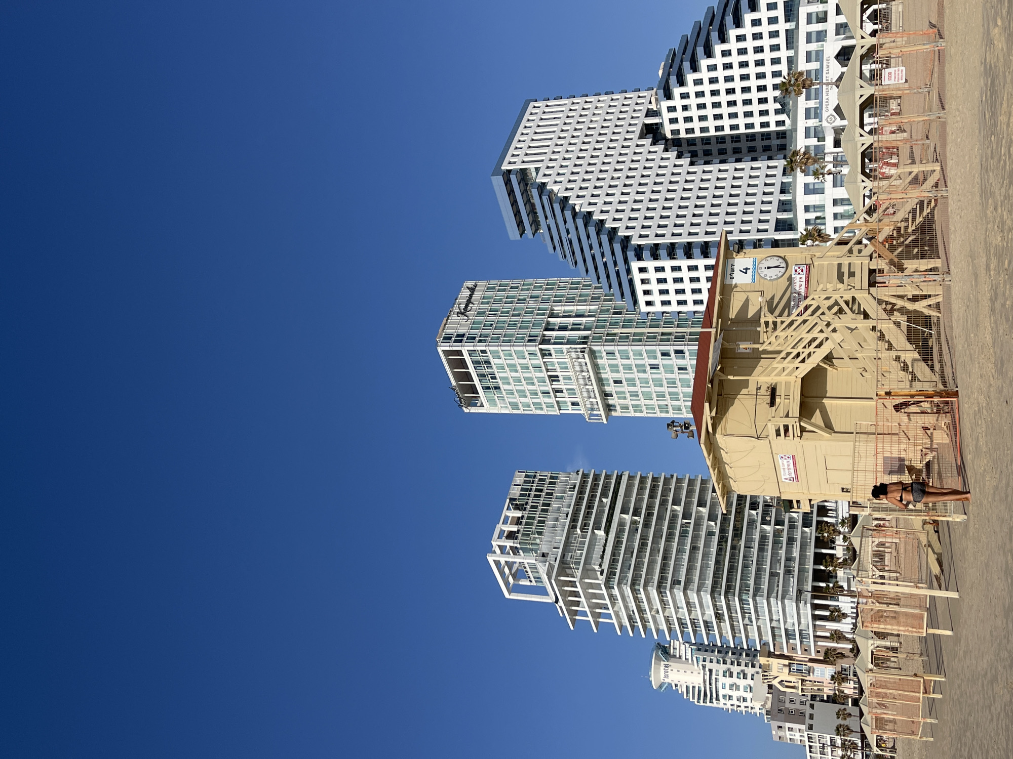 The image captures a beachfront scene in Tel Aviv, Israel, with modern buildings and a lifeguard station. The sky is bright blue, contrasting with the white and gray architecture of the buildings. A lifeguard station is present on the beach, with a woman nearby, seemingly exercising or stretching. The beach appears to be under some construction or preparation, as indicated by the presence of orange barriers and construction materials.
