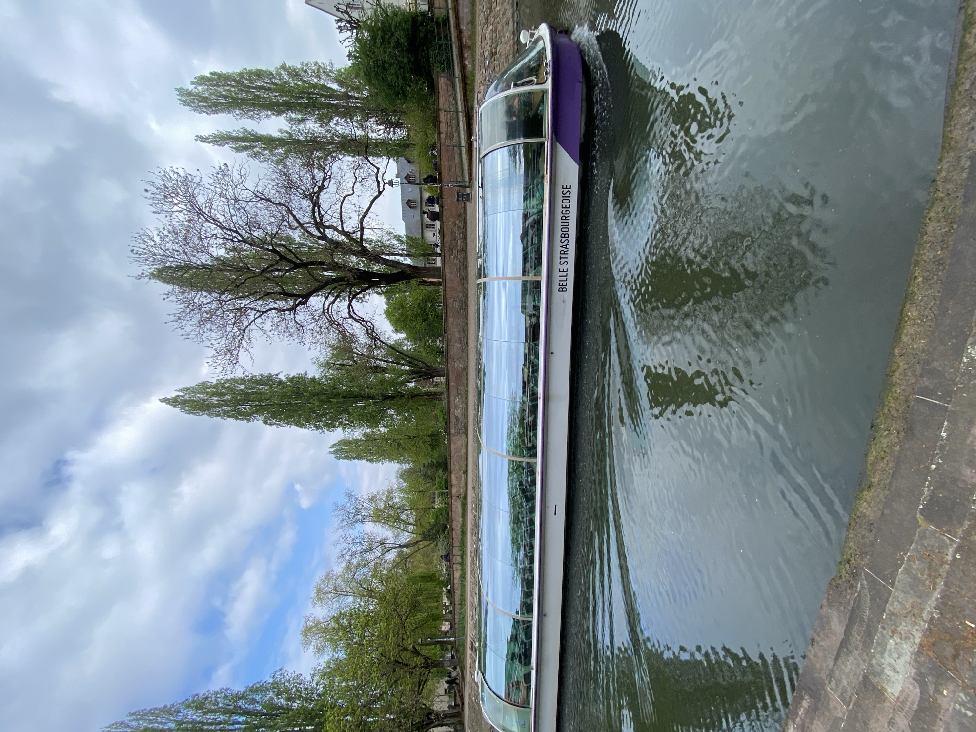 A full-view, medium shot captures a canal in Strasbourg, France, with a "Belle Strasbourgeoise" boat sailing on it.
The boat, primarily white with purple accents, travels along the canal’s murky, grey water. The boat's glass windows reflect the trees, buildings, and sky. "Belle Strasbourgeoise" is printed on the side of the boat in white lettering.
On the canal's left side, the stone bank is lined with various trees, ranging from budding spring foliage to stark, leafless branches. Tall, thin trees stand like sentinels beside a building with a grey roof and dormer windows.
The sky above features a mix of blue patches peeking through a thick blanket of grey and white clouds.