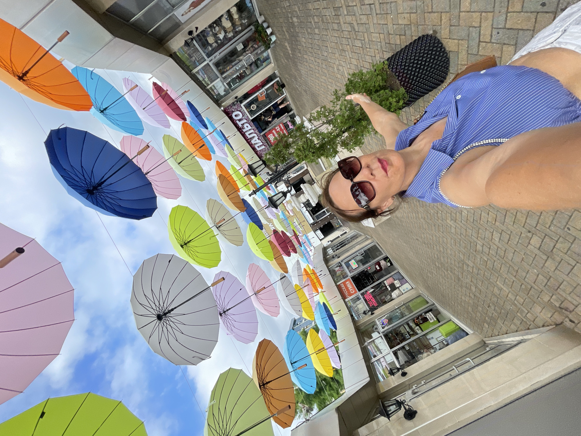 A woman with sunglasses and a blue striped shirt is taking a selfie outdoors. She is standing on a brick walkway in front of shops. Above her, many colorful umbrellas are suspended in the air. The sky is visible between the umbrellas. The scene appears to be a pedestrian street lined with businesses. The woman is pointing to her left with her right hand. The image is taken from a low angle, looking up at the umbrellas and the woman. The location is not explicitly stated to be Sochi, Russia, but it is possible given the style of architecture and the presence of Cyrillic text on shop signs.