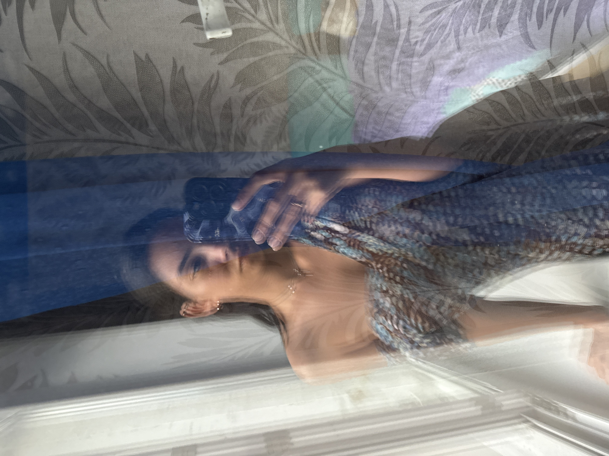 The image shows a woman with dark hair and fair skin, partially reflected in a window. She is holding a smartphone in her left hand. She wears a necklace and earrings. The reflection also shows part of a floral patterned wall or fabric. The lower part of the reflection seems to be the inside of a window. There is no discernible information about the location in the city of Chaikovsky, Russia.