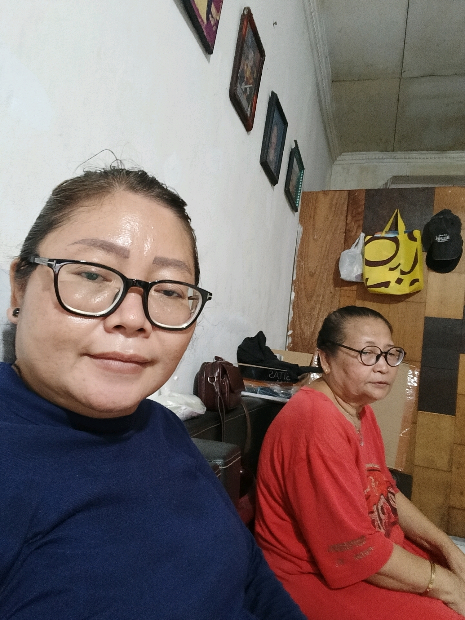 Two women are visible in this indoor, waist-up photograph. The woman in the foreground, on the left, is wearing a navy blue top and black-rimmed glasses. She has her hair pulled back in a bun and is looking directly at the camera with a slight smile. The woman in the background, on the right, is wearing a red shirt and round-rimmed glasses. She has her hair pulled back and is looking slightly to her left. The background consists of a white wall with several framed pictures hanging on it, and a wooden panelled wall with a yellow bag and a black cap hanging on it. The overall lighting suggests an indoor setting, possibly a home or a casual establishment, in Jatimulya, Indonesia.