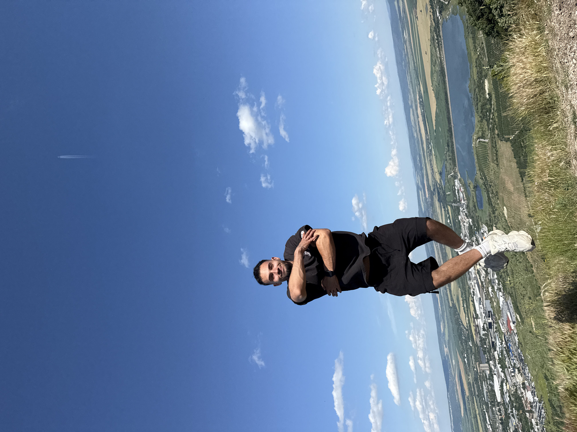 A man is pictured in the sky with his arms crossed and a smile on his face, wearing a black t-shirt and black shorts. He is standing on a grassy incline overlooking a city and a body of water. The sky is blue with scattered white clouds, and a plane can be seen in the distance. The image appears to have been taken on a clear, sunny day. The man's pose and expression suggest he is feeling confident and happy. The scene is a landscape with a city, fields, and a lake, indicating an outdoor location.