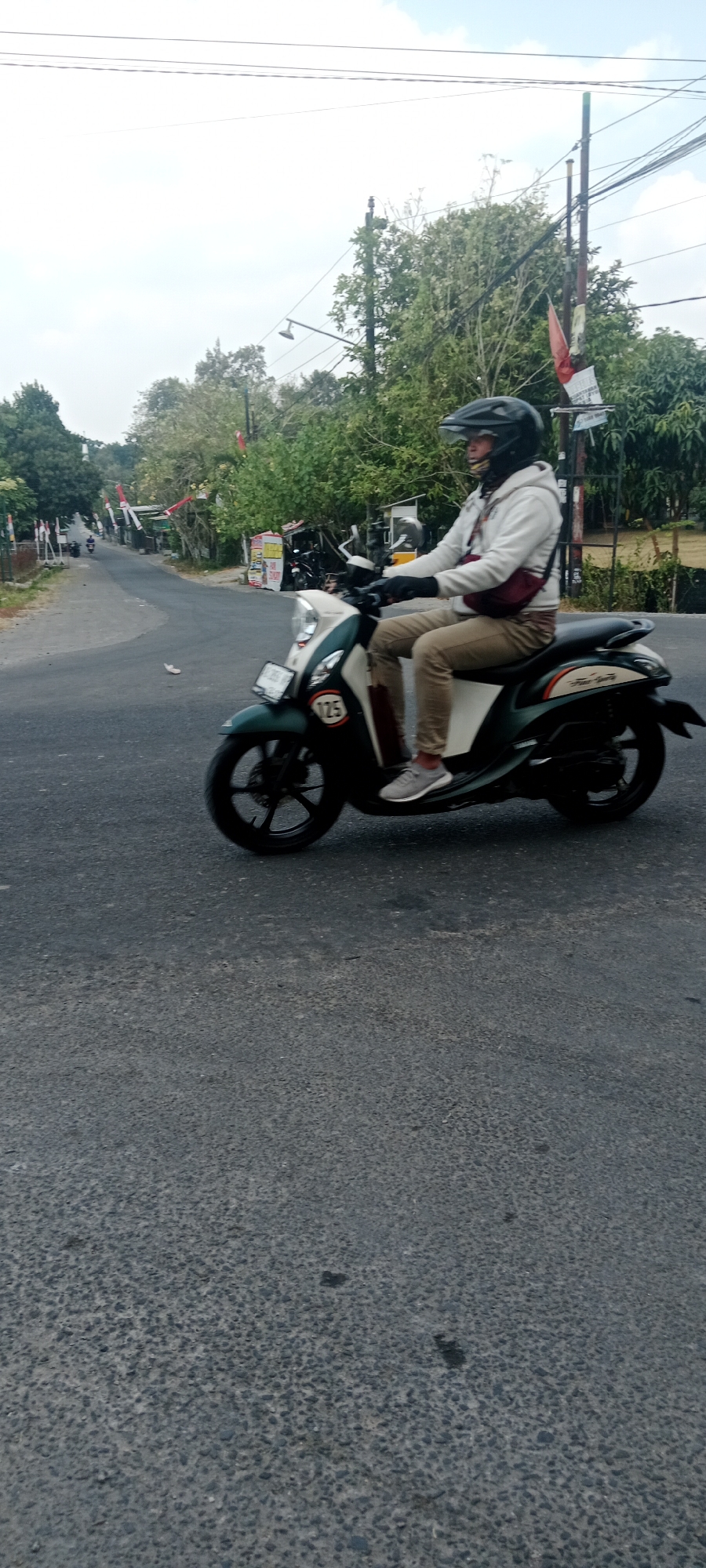 A person wearing a black helmet and a white hoodie is riding a dark green and white scooter on a paved road in Maguwoharjo, Indonesia. The person is wearing khaki pants and gray sneakers, and a dark red crossbody bag is slung over their left shoulder. The scooter has a white front fender and a dark green body with white accents. A circular emblem with the number "125" is visible on the front of the scooter. The road is gray asphalt and appears to be a two-lane street. On the right side of the road, there are trees and vegetation. In the background, there are several Indonesian flags and banners. The sky is overcast.