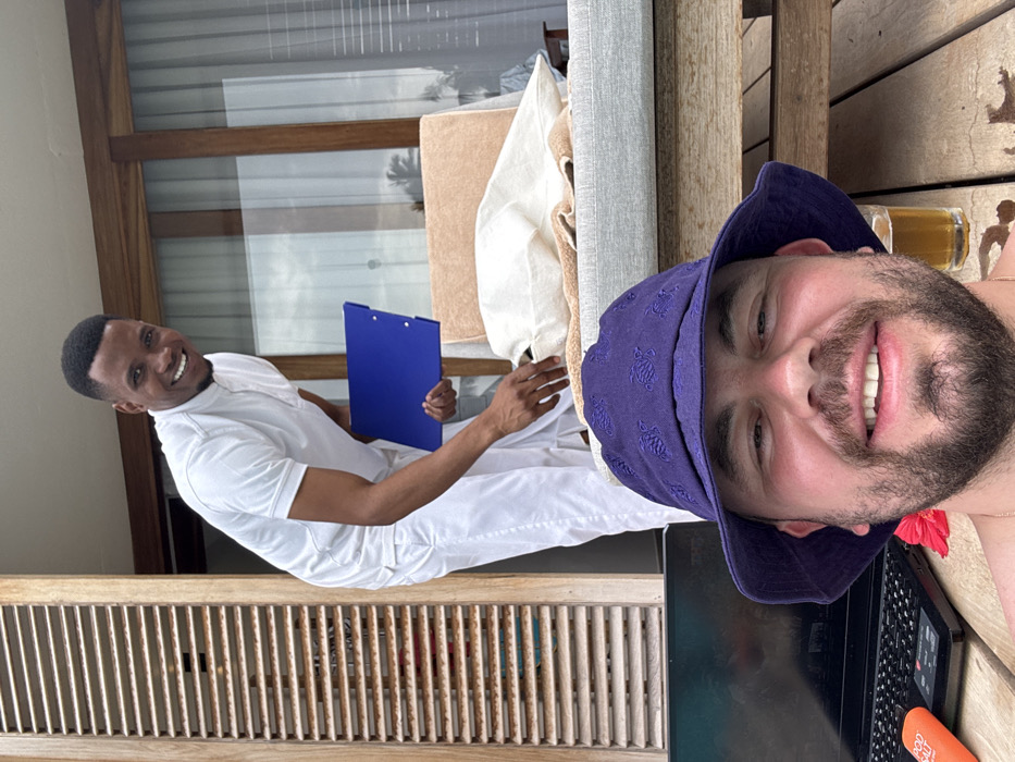 This image depicts two adult males on a wooden outdoor patio or balcony, likely at a resort in Zanzibar, Tanzania.
In the foreground, a Caucasian male with a beard and mustache, wearing a purple bucket hat with embroidered turtle patterns, is smiling directly at the camera, apparently taking a selfie. A red hibiscus flower is visible near his left shoulder. To his left, on the wooden decking, an open black laptop and an orange "POD SALT" branded bottle are visible. To his right, a glass containing an amber liquid is present.
In the mid-ground, an adult Black male, dressed in a white short-sleeved collared shirt and white trousers, is positioned behind and slightly to the right of the foreground subject. He is smiling and looking towards the camera, holding a blue clipboard in his left hand. His right hand is reaching towards a light grey cushioned lounge chair, which is covered with a beige towel and a white cloth.
The background features large glass panels framed by wooden structures, revealing lush tropical vegetation, including palm trees, under an overcast sky. To the far left, a section of wooden louvered shutters is visible. The scene suggests a relaxed, leisurely setting with a service interaction occurring between a guest and a staff member.