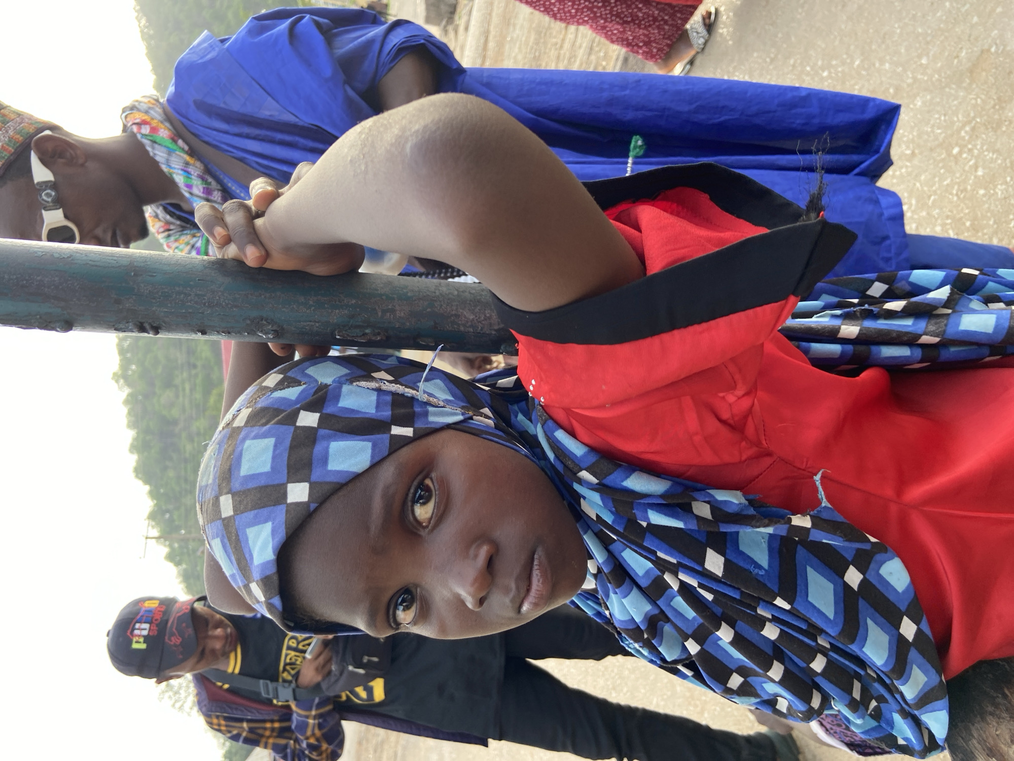 This close-up, portrait-oriented image, rotated 90 degrees clockwise, depicts several individuals gathered outdoors in Mararaba, Nigeria, during the daytime.
The primary subject is a young person with dark skin and dark eyes, positioned in the foreground and looking directly at the camera with a calm, neutral expression. They wear a red top with black trim, and a head covering featuring a striking blue, black, and white geometric pattern. Their arm rests on a dark metal railing that bisects the frame.
To the left, another person, possibly a young man, also leans on the same railing. He has dark skin, is wearing a blue garment over a colorful, plaid-patterned scarf, and has a decorative white ear cuff or earring visible. A textured cap is on his head, and his head is slightly bowed.
In the background, a person wearing a dark cap with partially visible colorful text, "PUMA SPORT," can be seen looking downwards. Further back and to the right, a person's legs and feet, clad in a red patterned garment and sandals, are partially visible. The ground appears to be a light-colored paved or concrete surface. Beyond the immediate gathering, the background shows lush green foliage and bright, possibly hazy, daylight sky, suggesting a natural outdoor setting. The light quality indicates it is daytime.