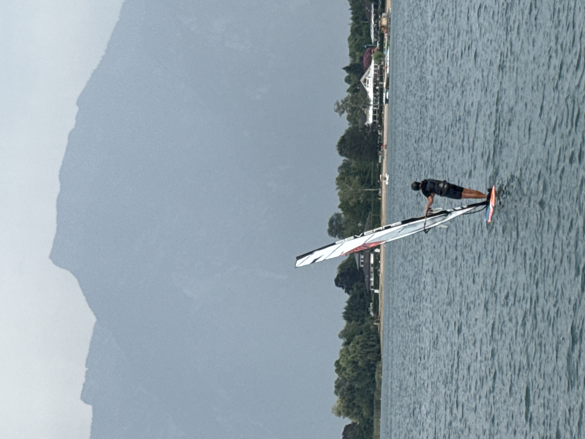 A person is windsurfing on a body of water. The individual is wearing a black helmet and a black vest over dark clothing, and is standing on a windsurf board with a sail. The water appears somewhat choppy. In the background, there is a shoreline with trees and buildings, and behind that, large mountains. The sky is overcast and gray. The location is Calceranica al Lago, Italy.