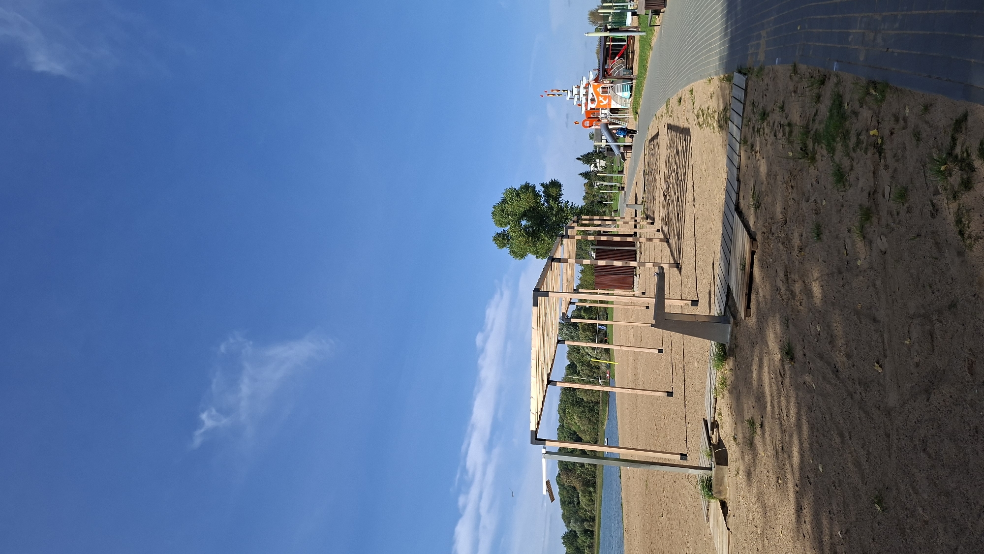 A beach and park area is depicted on a bright, clear day with a blue sky and scattered white clouds. In the foreground, a sandy beach is visible with wooden plank walkways and a shaded pergola structure. Beyond the beach, a body of water, likely a lake, is present, bordered by a line of trees. To the right of the scene, a paved walkway curves away, lined with benches and a children's playground designed to resemble a ship. A person is walking on the paved path. The playground features slides and climbing structures. The overall environment suggests a recreational space.